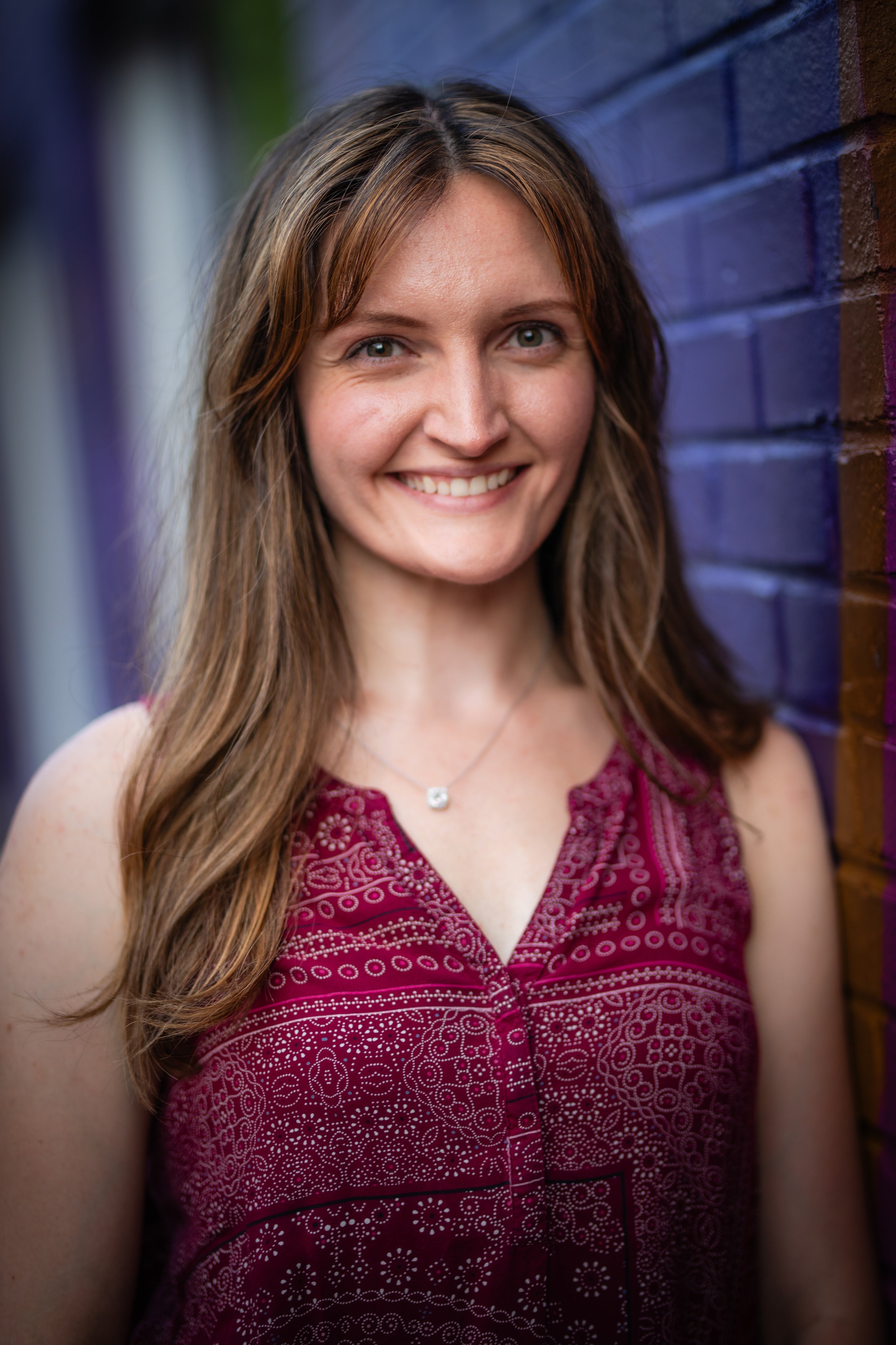 Portrait of a woman with long brown hair smiling, wearing a red patterned sleeveless top, standing next to a dark purple and brown brick wall.