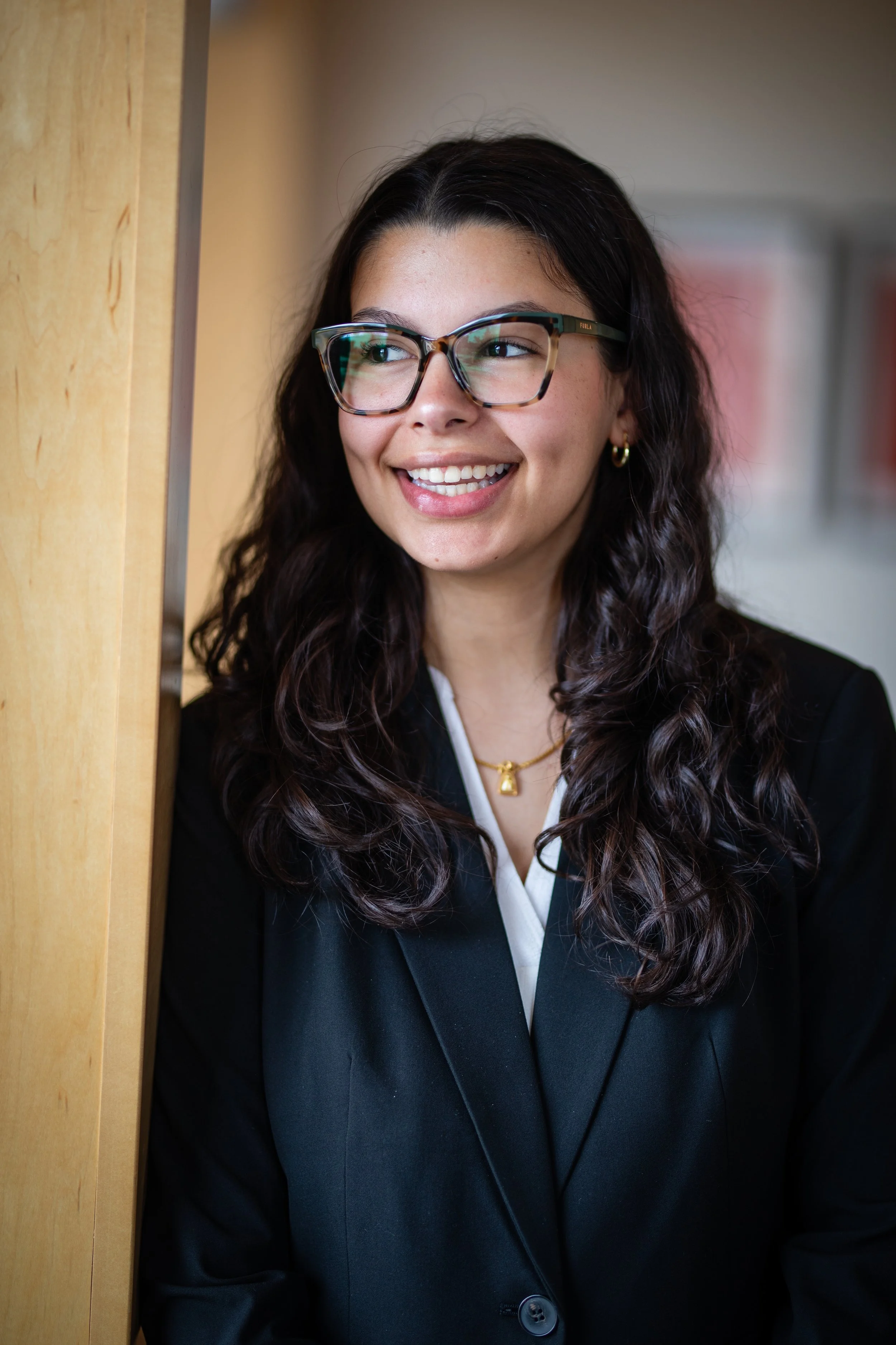 A young woman with curly dark hair, wearing glasses, a black blazer, and gold jewelry, smiling and leaning against a wooden surface.