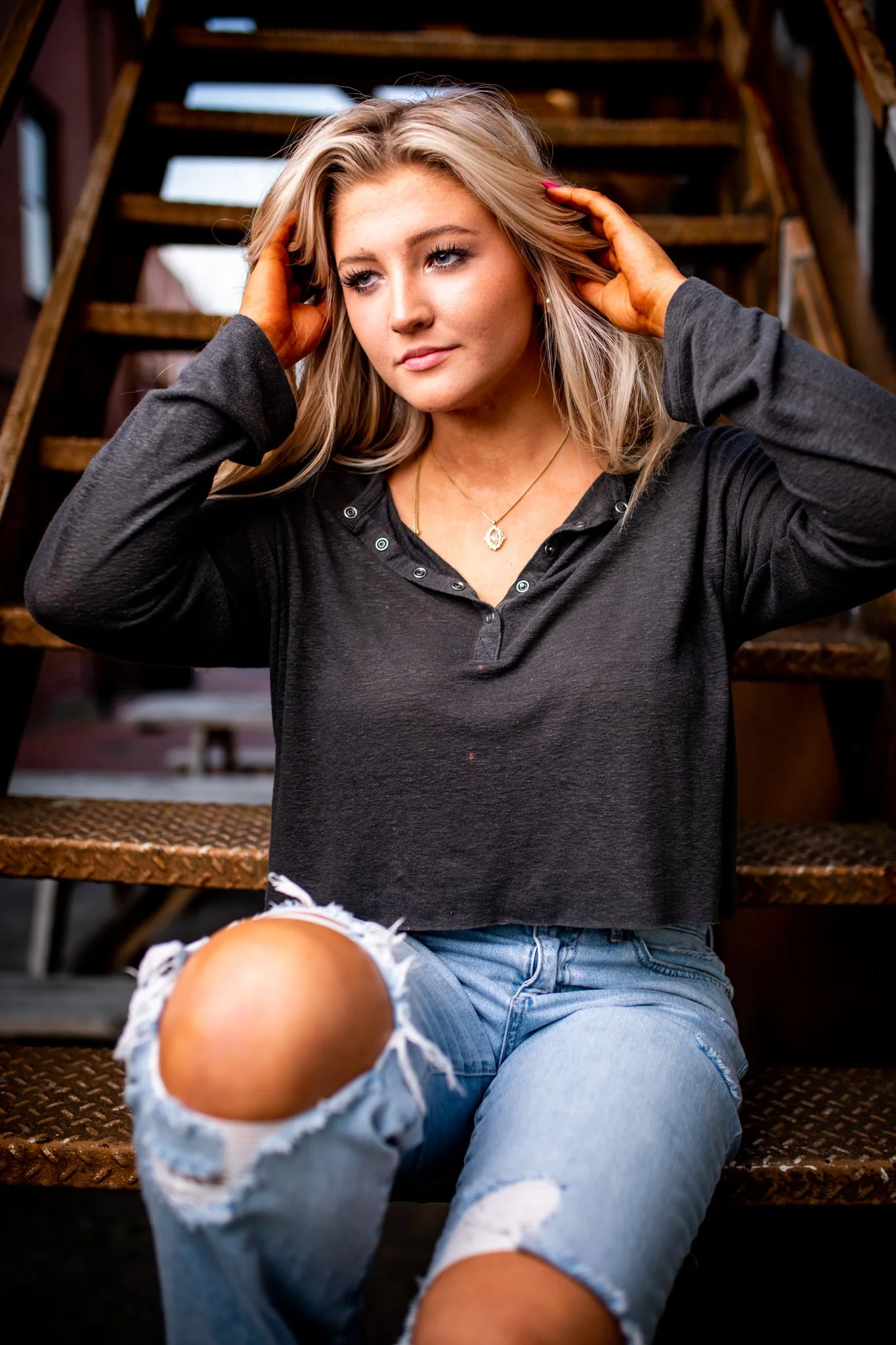 A young woman with blonde hair sitting on metal stairs adjusting her hair, wearing a black long-sleeve shirt and ripped jeans.