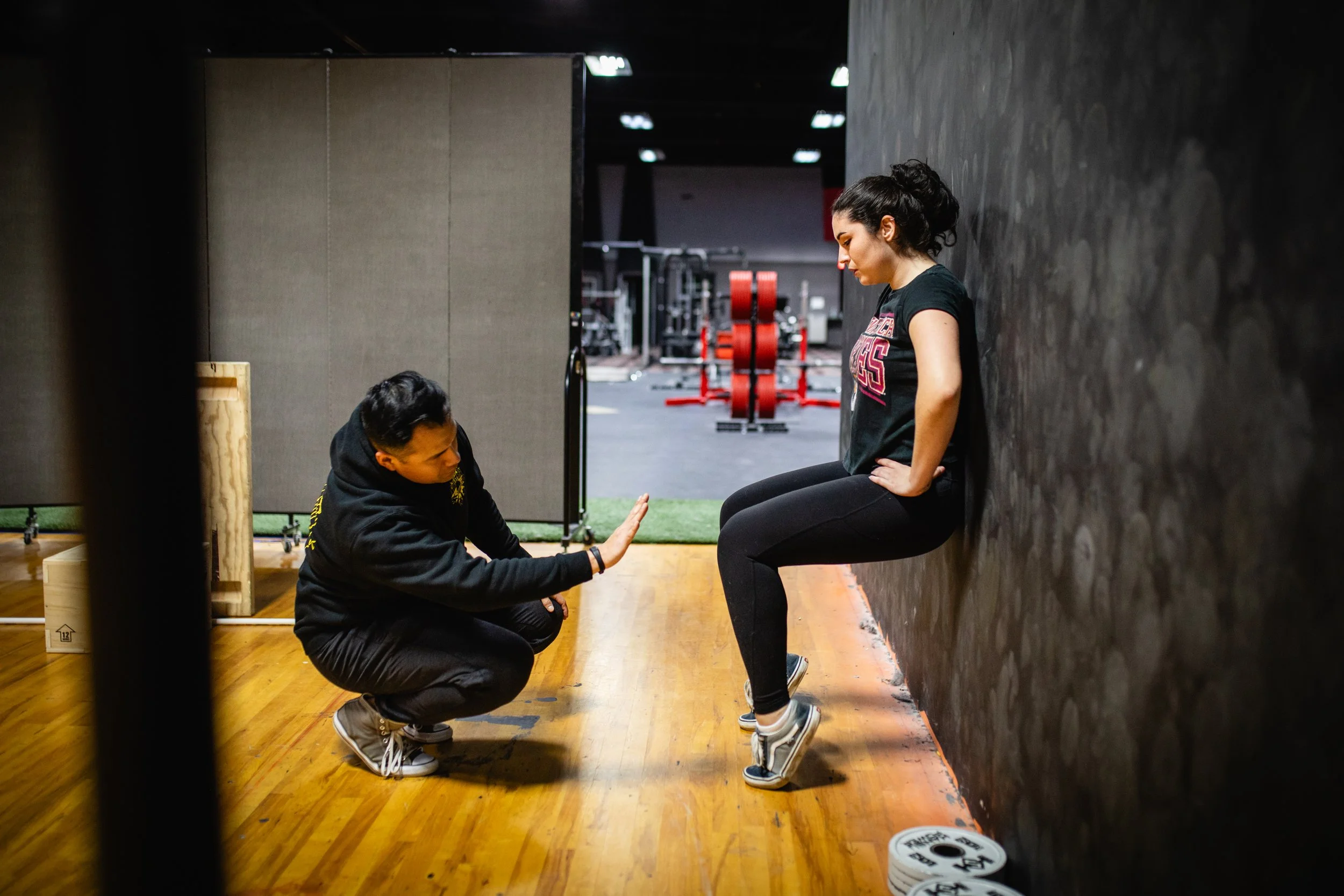 A personal trainer crouching in front of a woman doing wall squats in a gym. The woman wears a black t-shirt and black leggings, with her hands on her hips, while the trainer gives directions or encouragement.