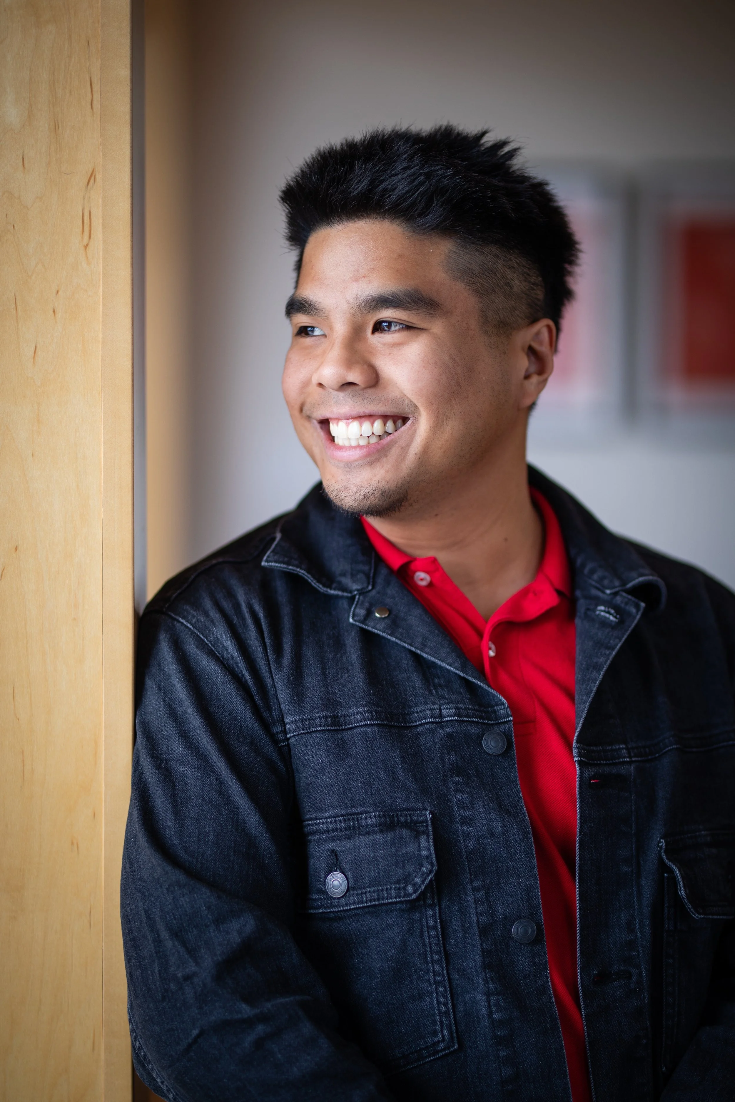 A young man with short, black spiked hair and a goatee, smiling while leaning against a wooden wall.