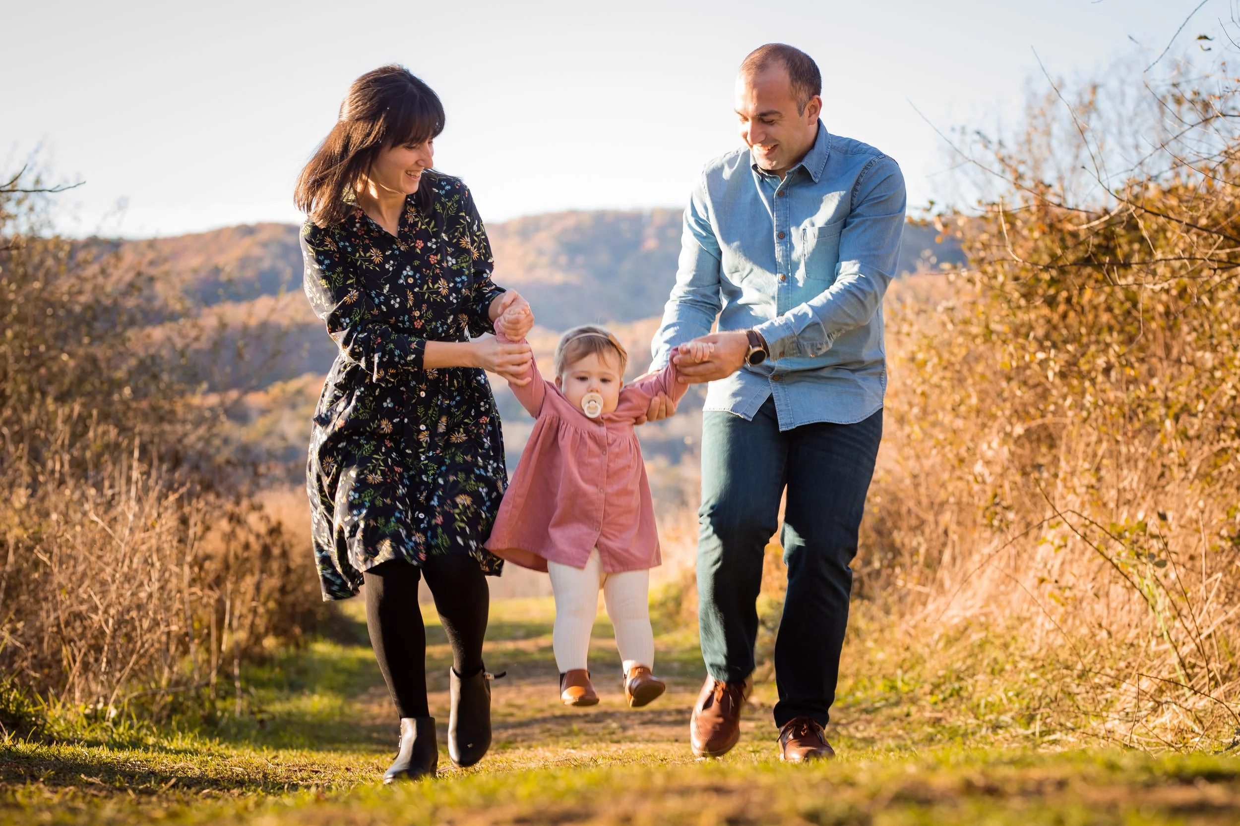 A family of three, a woman, a man, and a young girl, enjoying a walk outdoors in autumn, with the father and mother holding the daughter's hands as she is lifted off the ground.