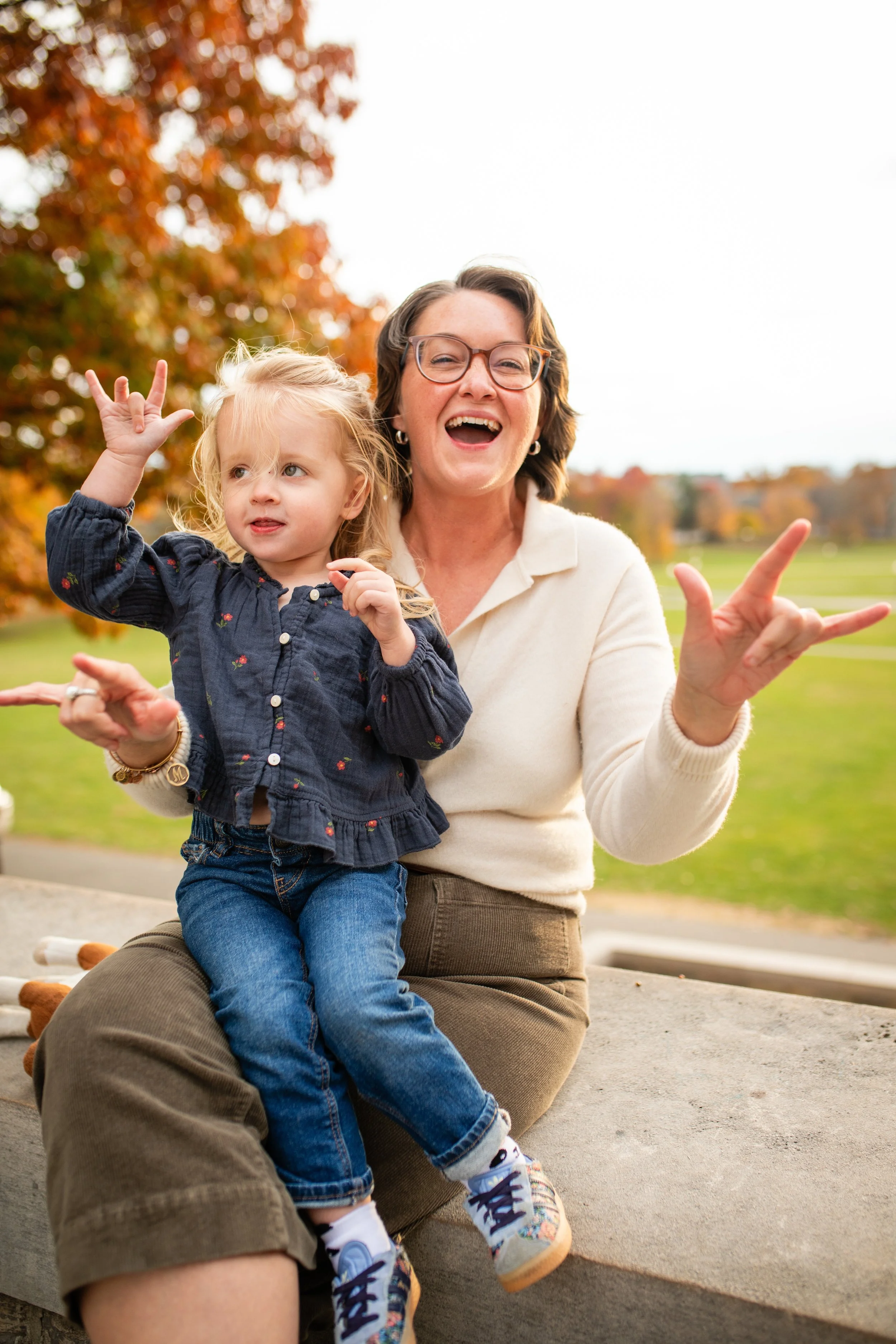 A woman and a young girl sitting outdoors on a concrete ledge, smiling and making hand gestures. The background features autumn trees with orange leaves and a green park.