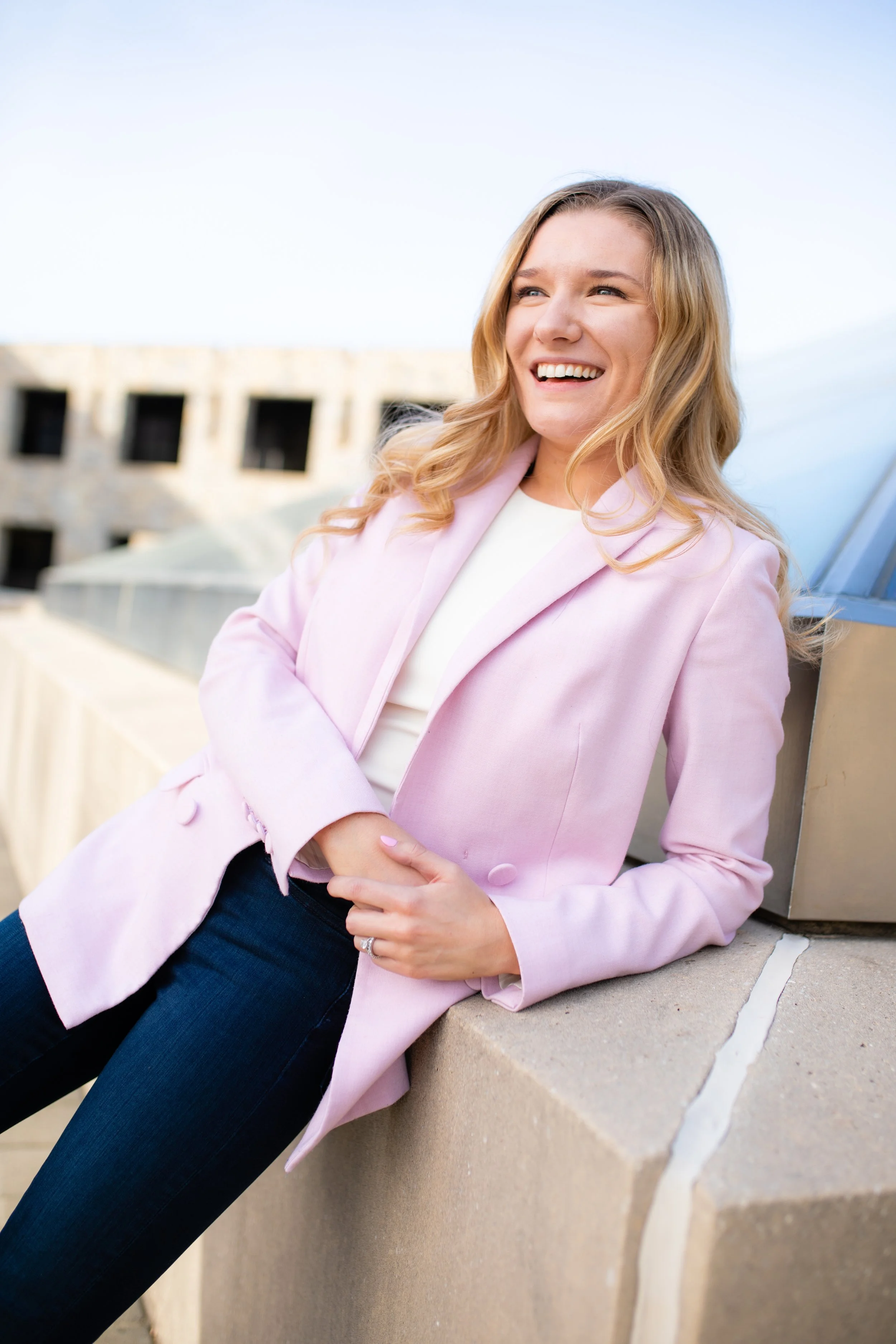 A woman with blonde hair wearing a pink blazer and dark jeans is smiling and leaning against a concrete ledge on a rooftop.