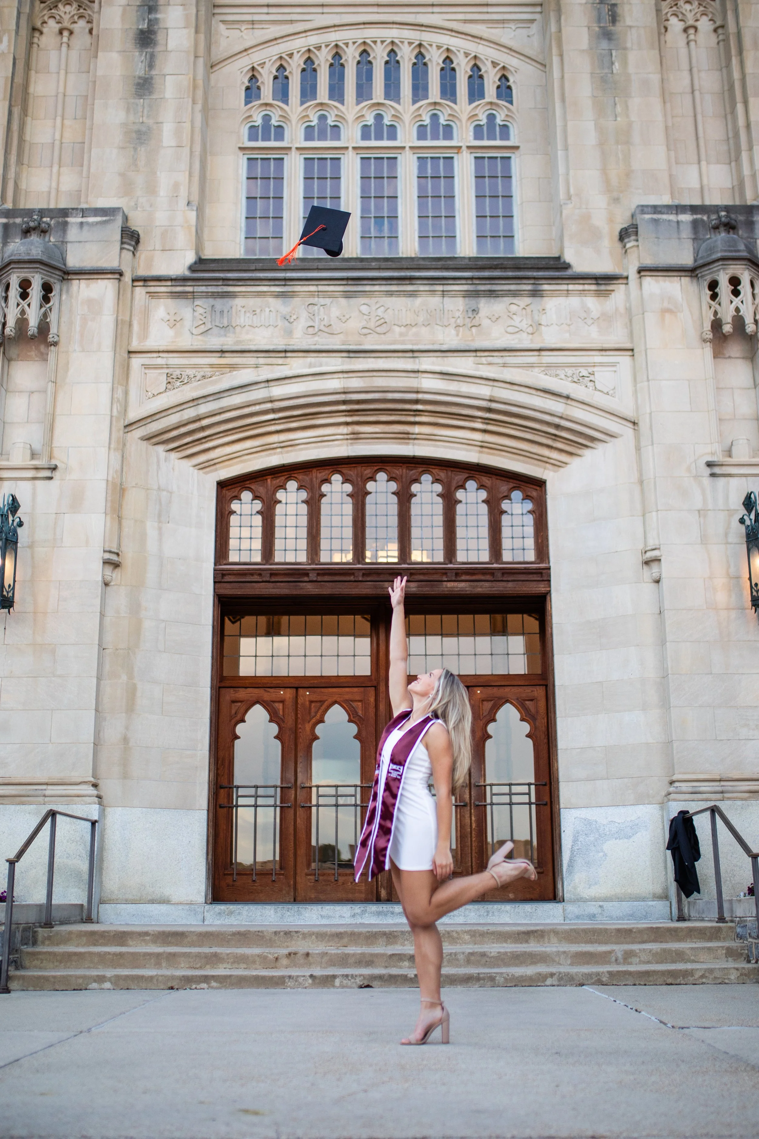 A woman in a white dress and heels poses as she throws a graduation cap into the air outside a large stone college building with arched windows, during the evening.