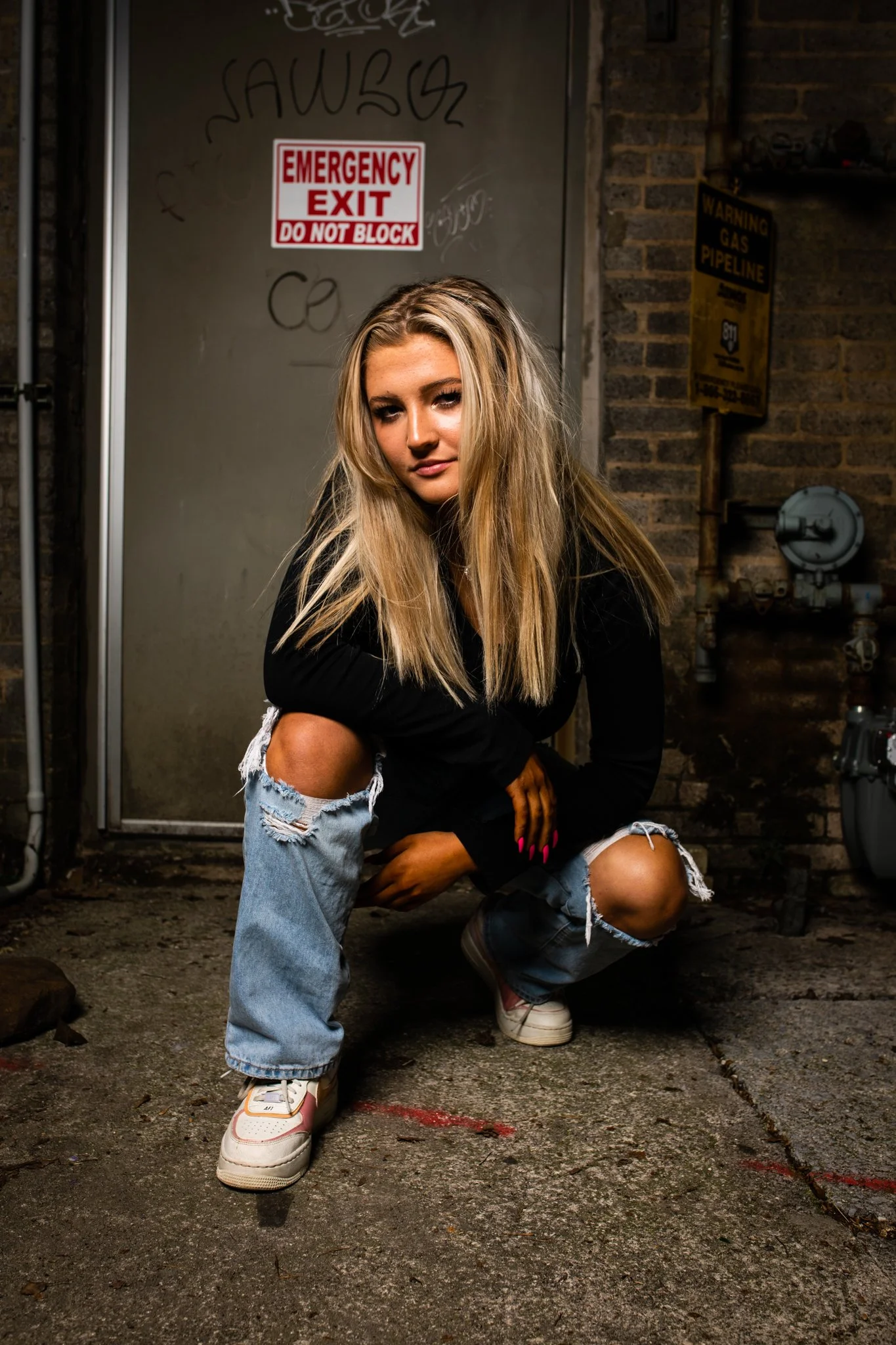 A young woman with long blonde hair, wearing a black long sleeve shirt and ripped jeans, squatting on a concrete sidewalk in front of a metal door and brick wall, with signs that read 'Emergency Exit Do Not Block' and 'Warning Gas Pipeline' behind he