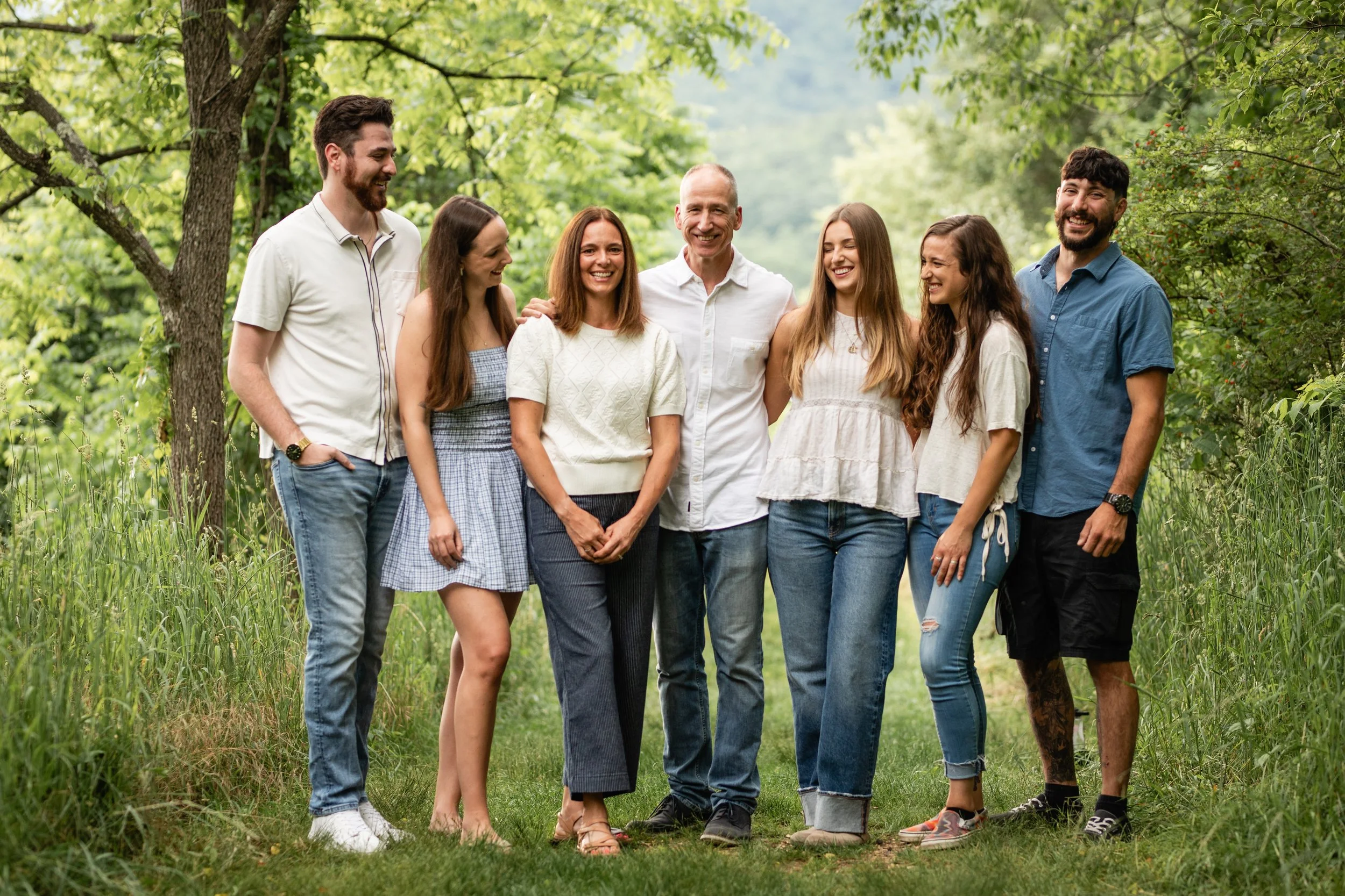 Group of seven people standing outdoors on green grass, smiling and enjoying each other's company, surrounded by trees and greenery.