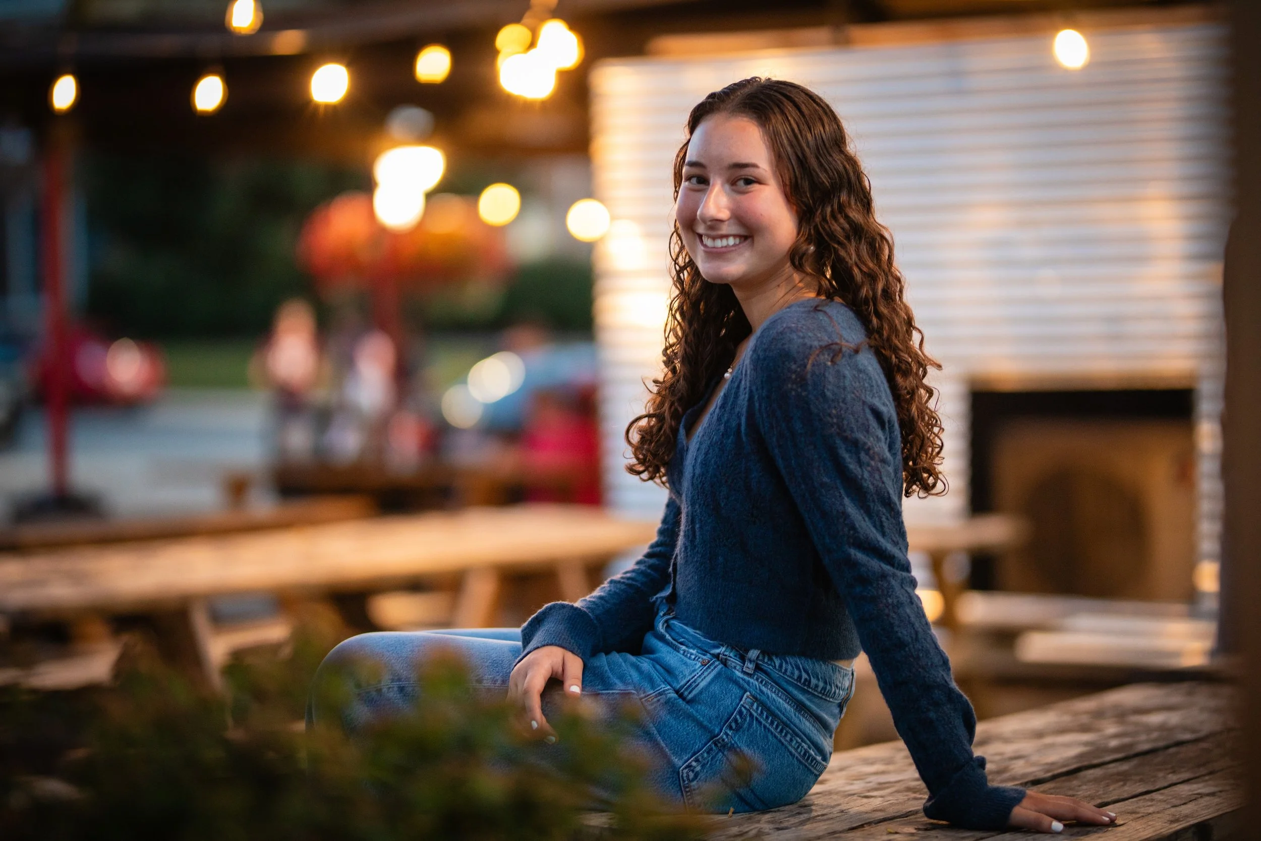 A young woman with curly brown hair, wearing a dark blue sweater and blue jeans, sitting on a wooden bench at an outdoor area during evening, smiling at the camera with string lights glowing in the background.