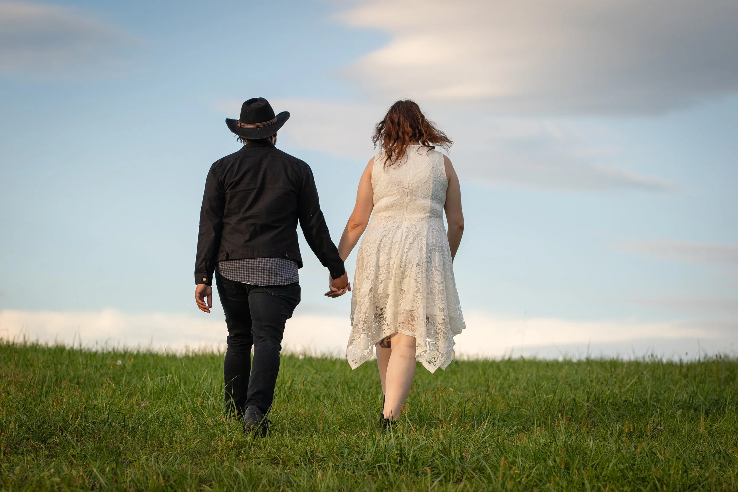 A couple walking hand in hand across a grassy field under a cloudy sky, the woman wearing a white lace dress and the man in dark clothing with a black hat.