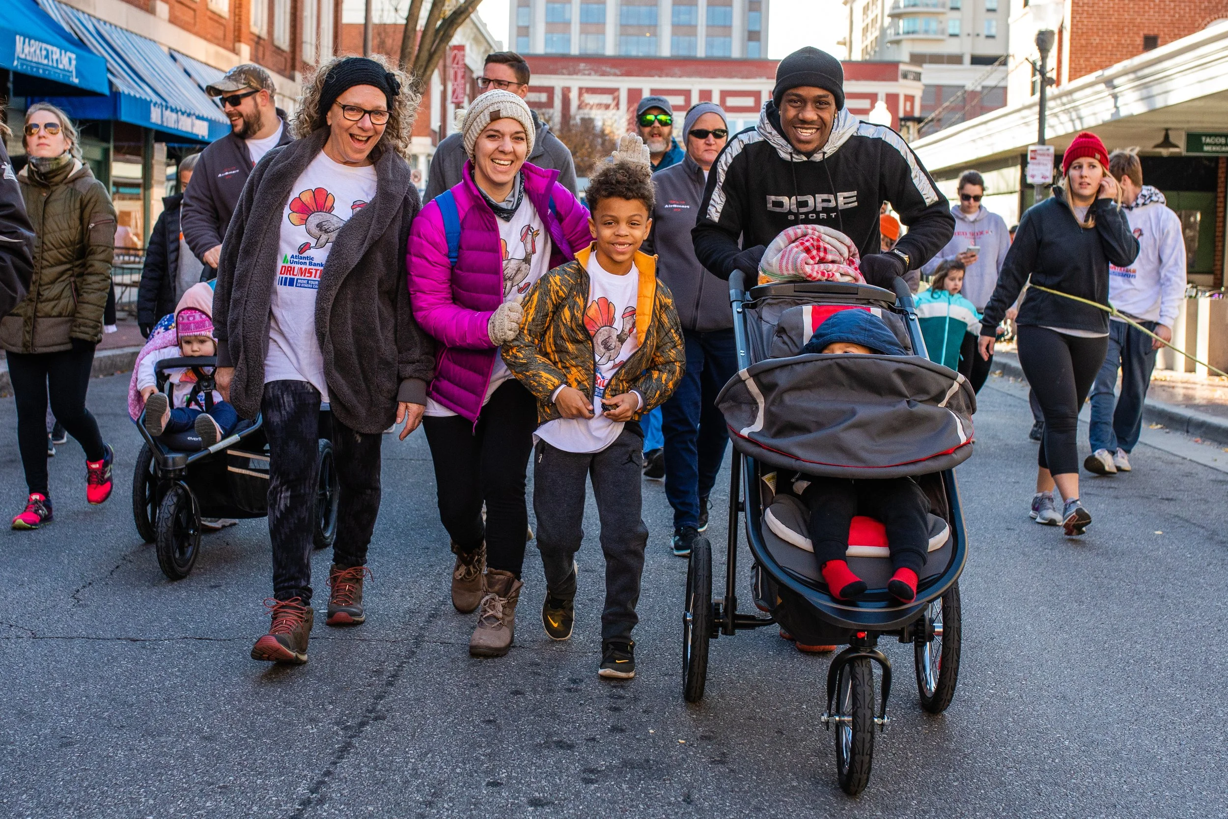 A diverse group of people walking on a city street during a parade or event. Some are pushing strollers with children, smiling, and enjoying the day. The group includes women, children, and men dressed in casual winter clothing.