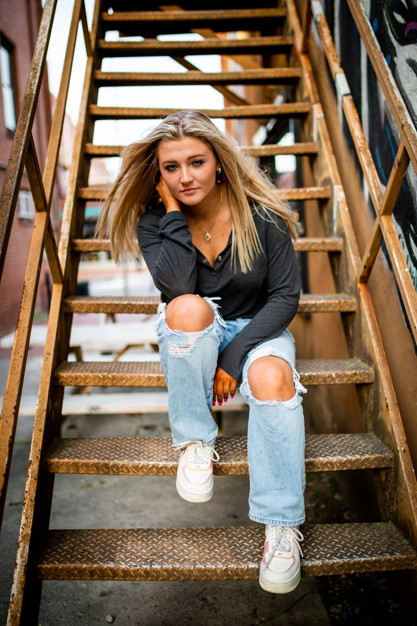 A young woman with blonde hair, wearing a black long-sleeve shirt, ripped light blue jeans, and white sneakers, sitting on a metal staircase outdoors.