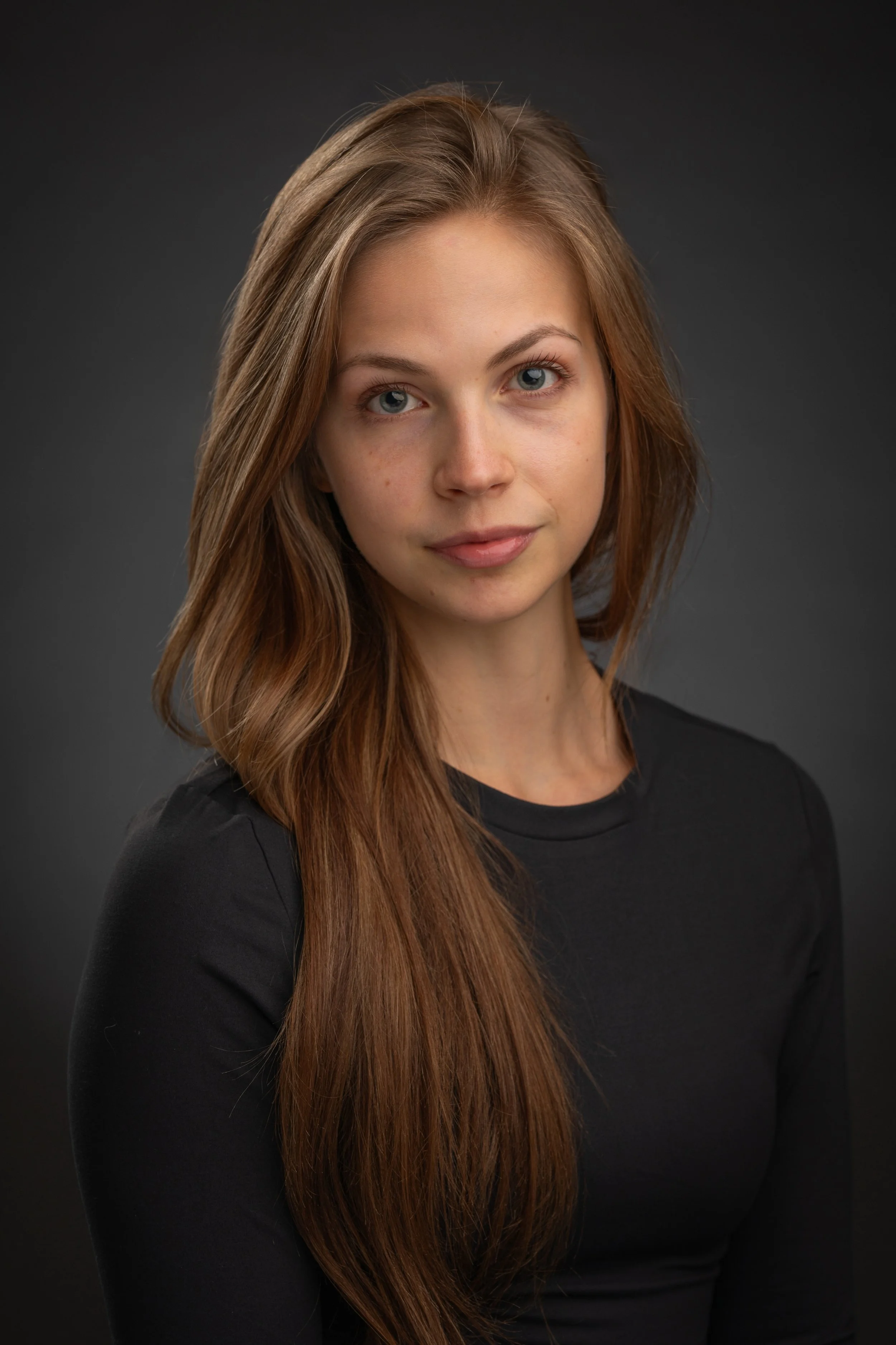 A woman with long reddish-brown hair, blue eyes, and fair skin wearing a black shirt against a dark gray background.