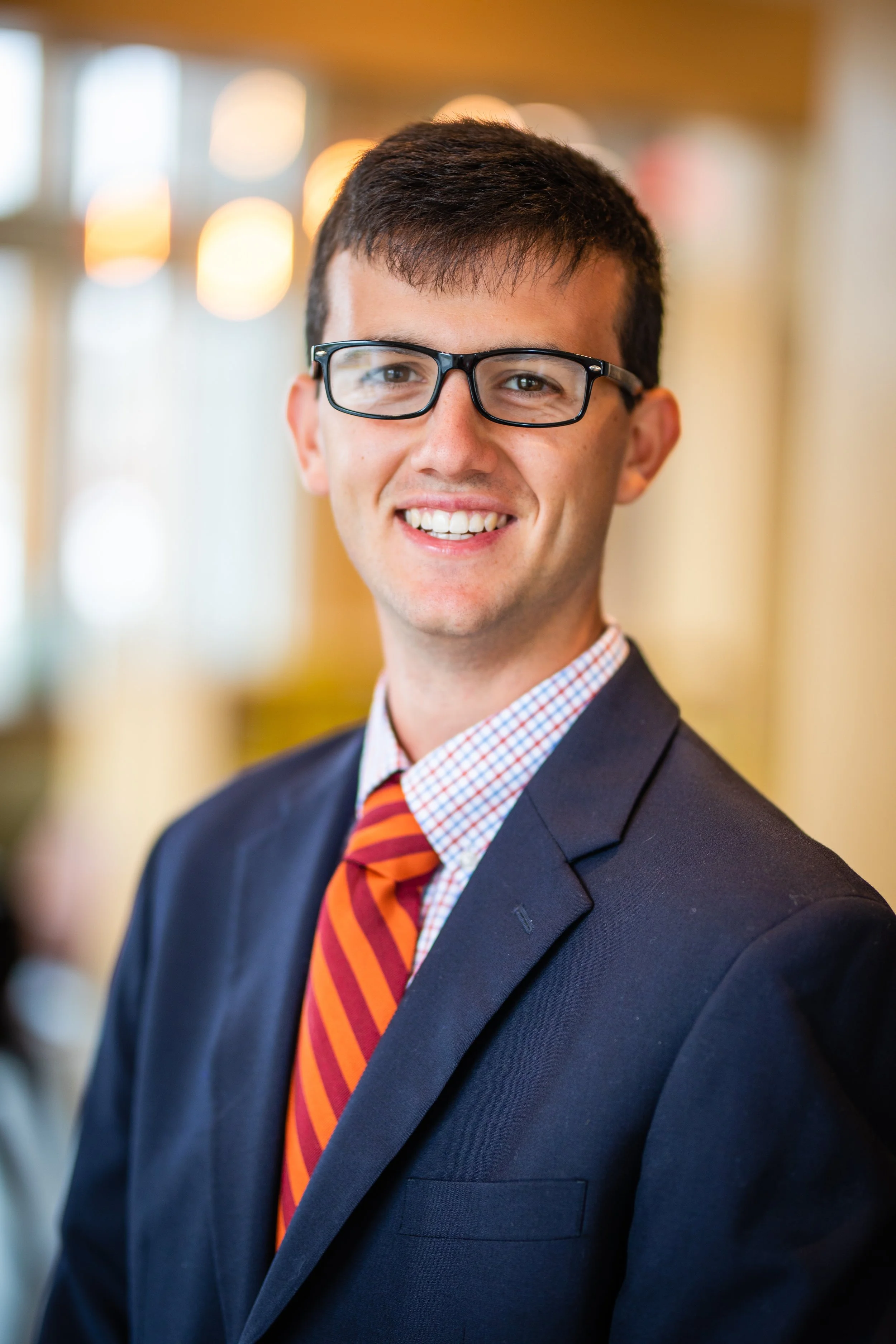Headshot of a smiling young man wearing black glasses, a dark suit, a light checkered shirt, and a red and orange striped tie, in a brightly lit indoor setting.