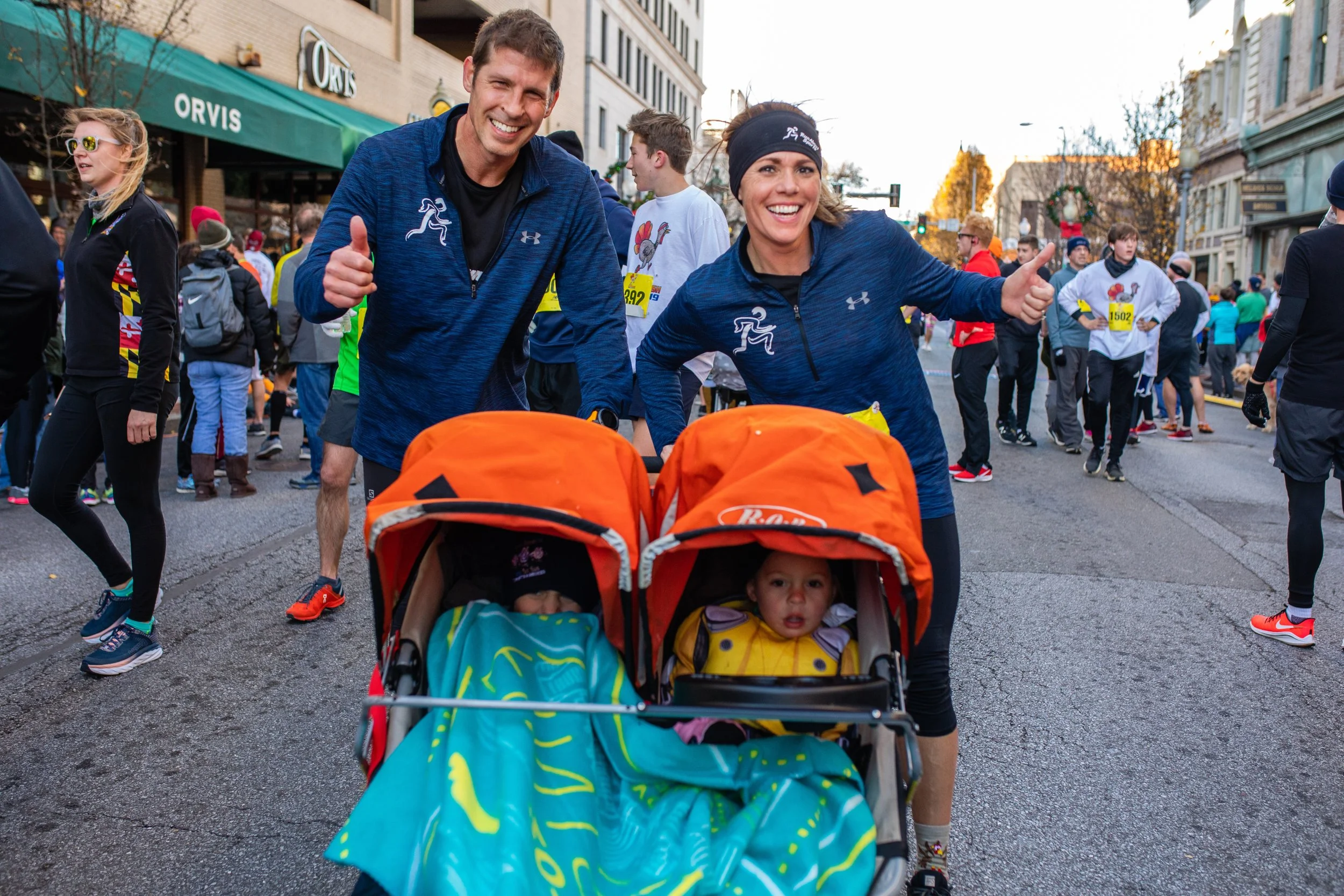 A smiling man and woman pushing strollers with two young children during a marathon or race event on a city street filled with runners and spectators.