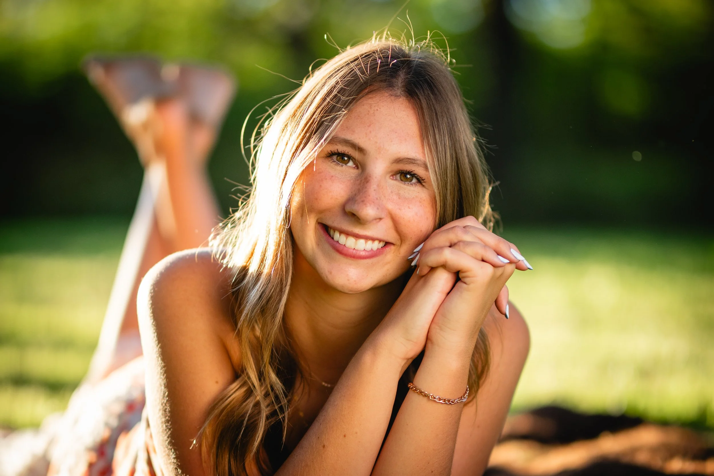 A young woman with long, wavy hair lying on her stomach outdoors on green grass, smiling and resting her chin on her clasped hands, with blurred green foliage in the background.