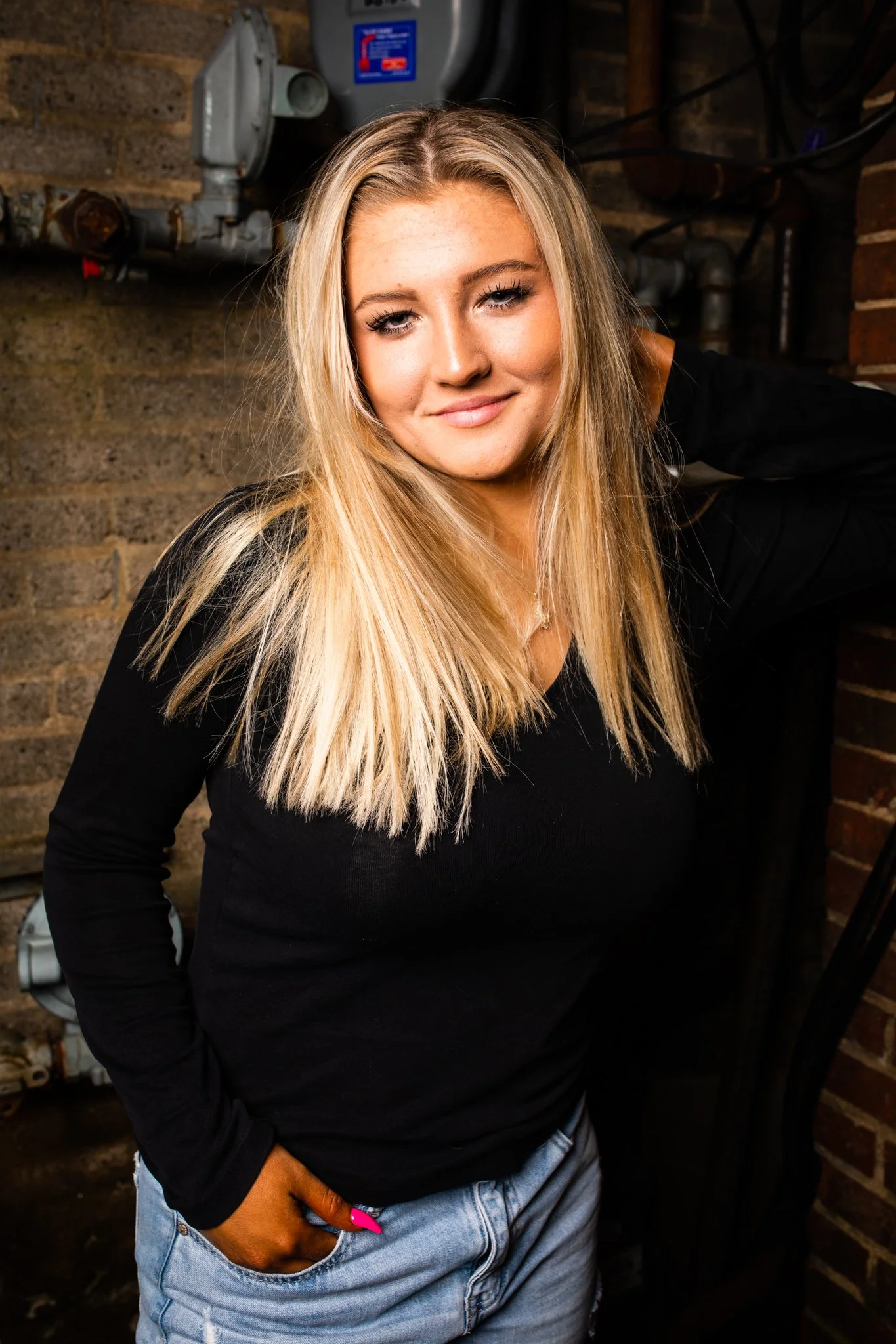 A young woman with long blonde hair, wearing a black top and light blue jeans, standing against a brick and concrete wall with pipes visible in the background.