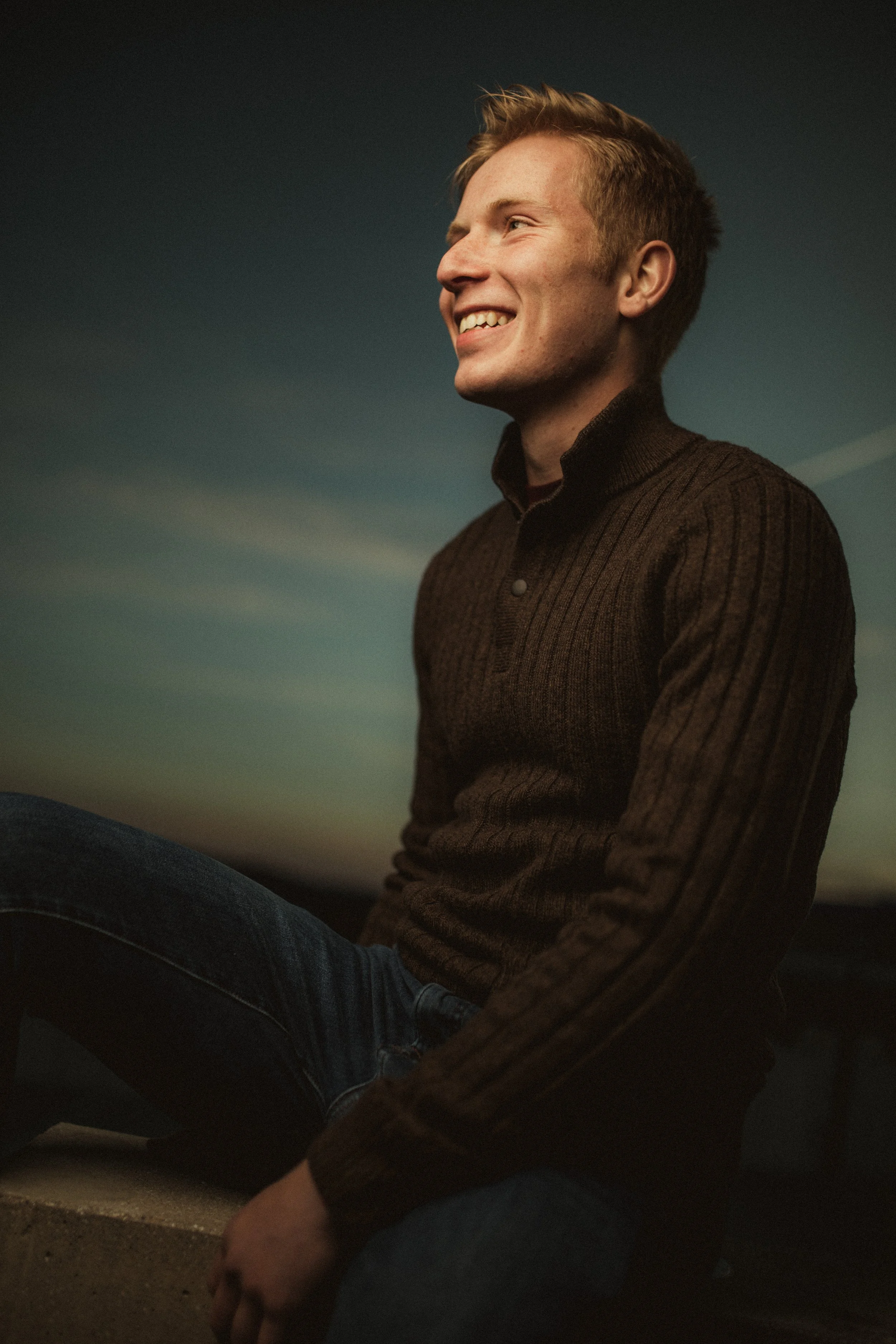 Young man with short blonde hair, wearing a dark brown sweater, sitting outdoors during sunset, smiling and looking to the right.