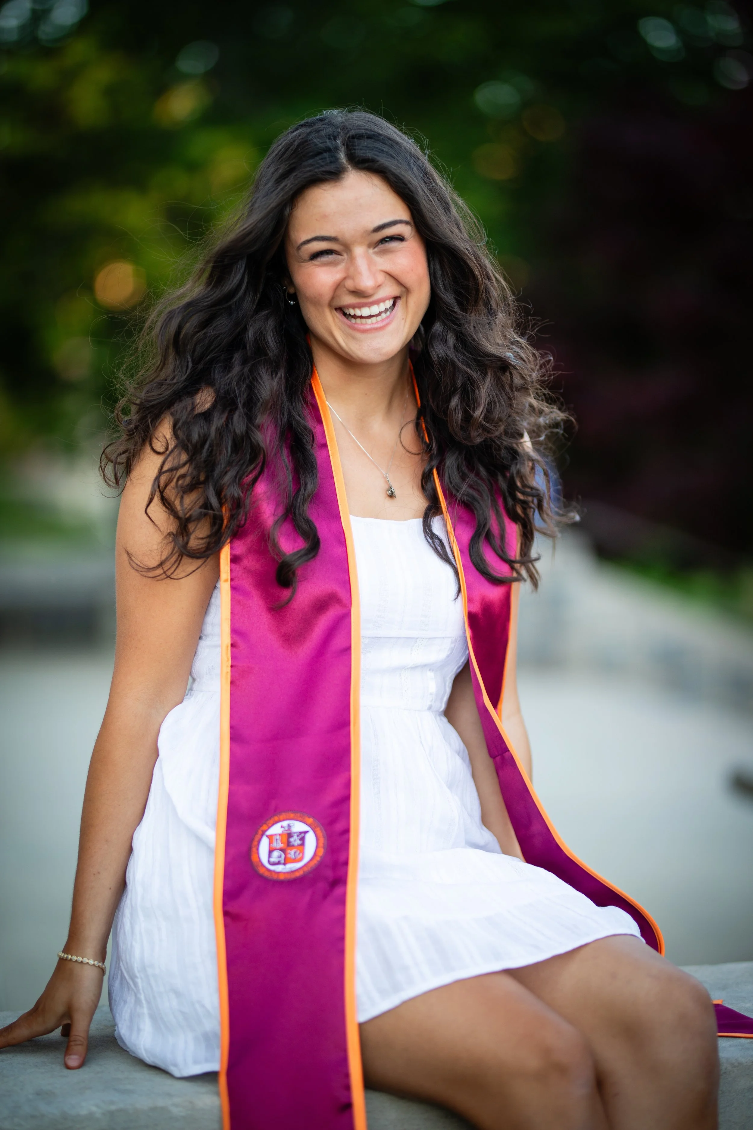 Young woman in white dress wearing a maroon and orange graduation stole, smiling outdoors.