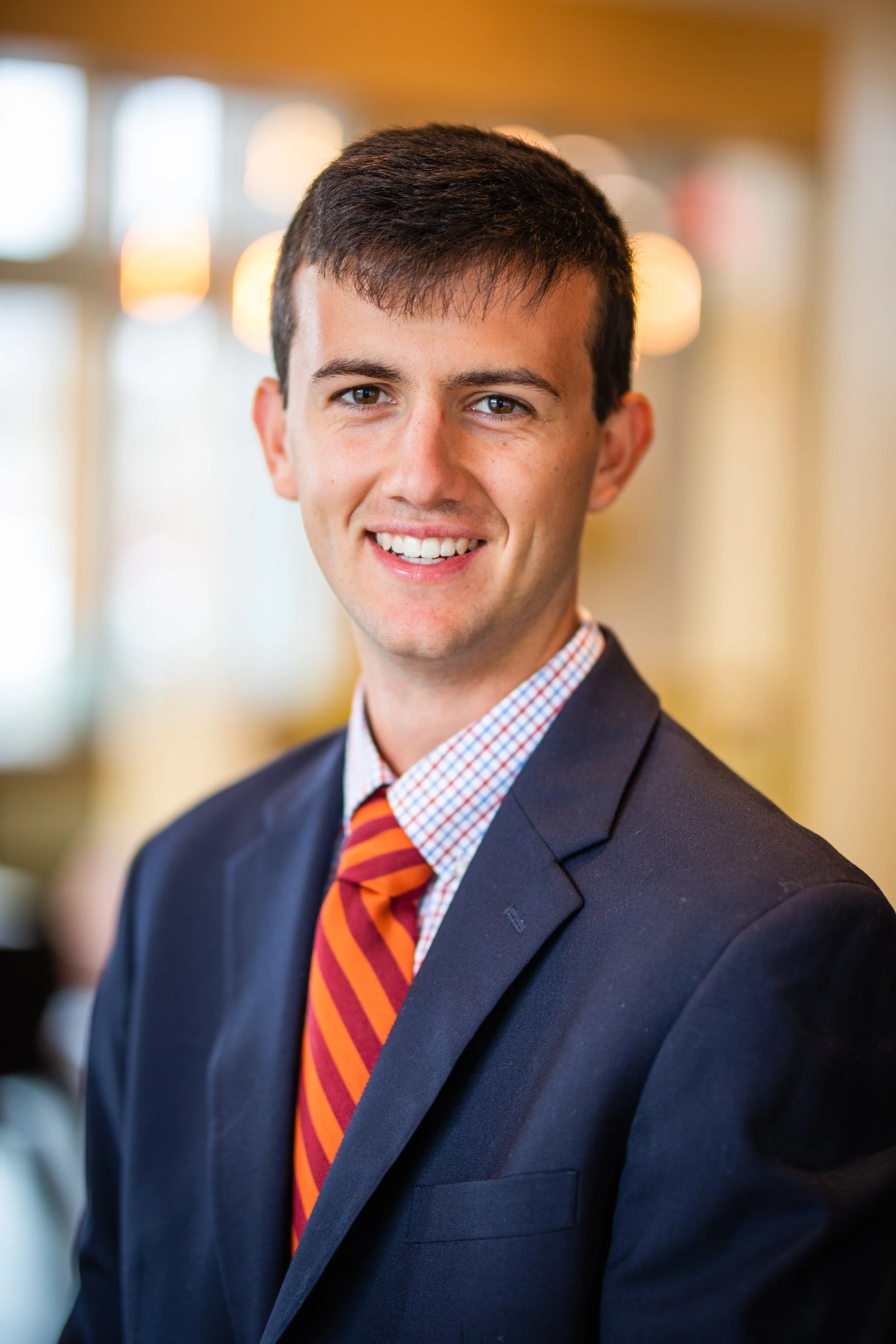 A young man wearing a dark suit jacket, a checkered dress shirt, and a red and orange striped tie, smiling at the camera in a bright indoor setting with blurred warm lighting in the background.