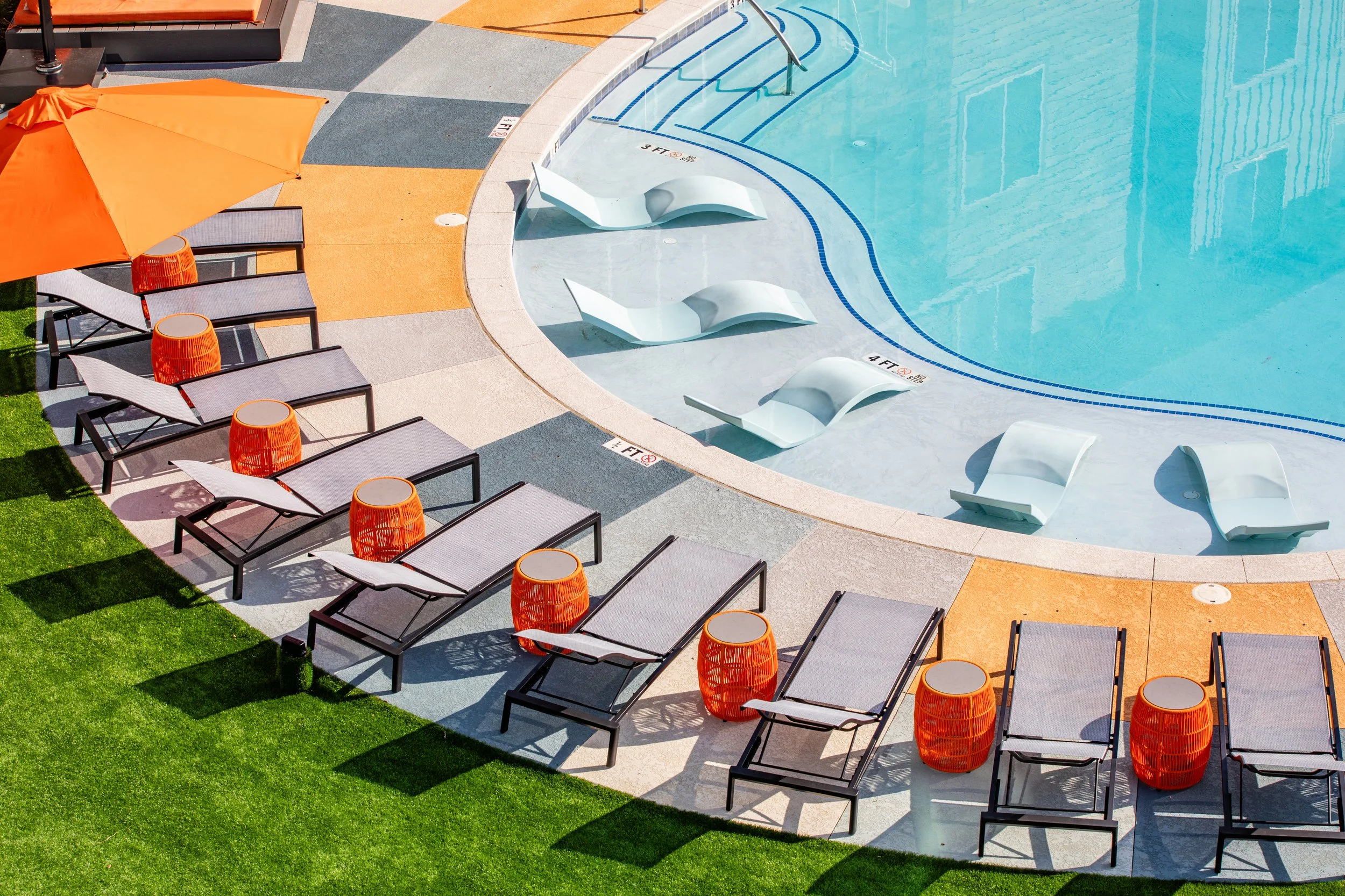 Empty poolside area with lounge chairs, orange side tables, and an orange umbrella, next to a swimming pool.