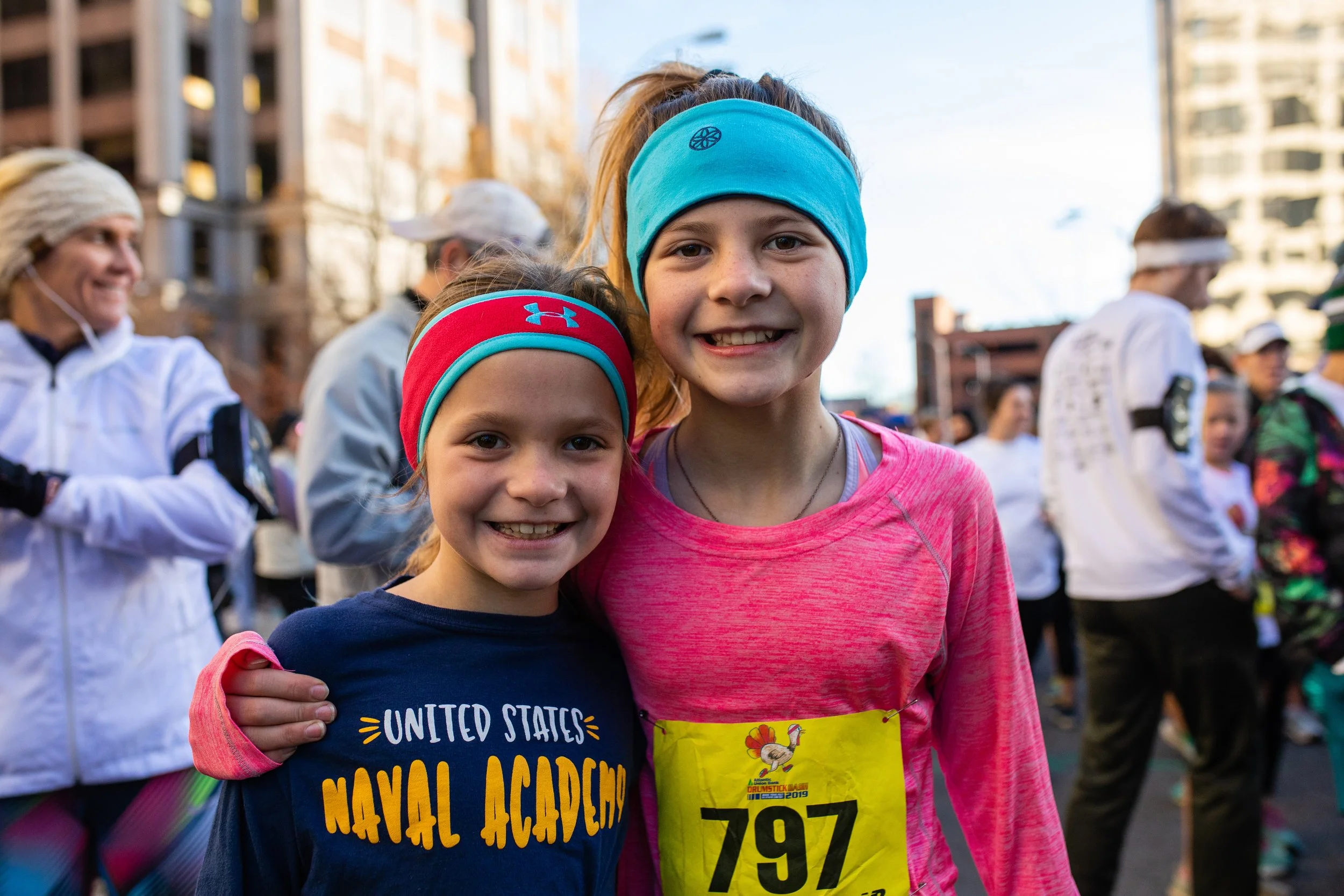Two young girls smiling and standing close together at a race event, with one wearing a navy shirt that reads 'United States Naval Academy' and a red headband, and the other wearing a pink long-sleeve shirt with a yellow bib labeled '797'. The backgr
