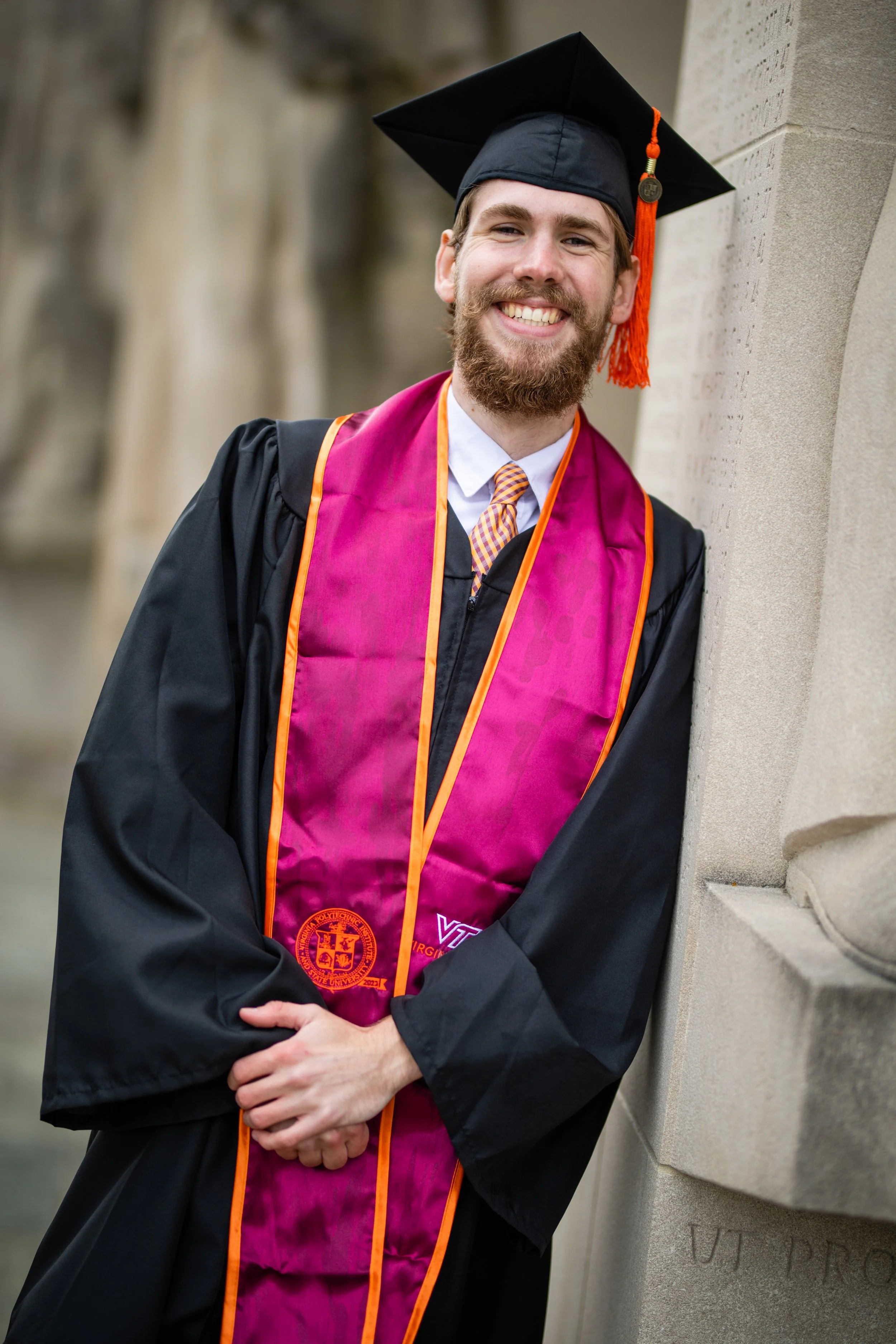 Graduating man in cap and gown smiling and leaning against a stone wall.