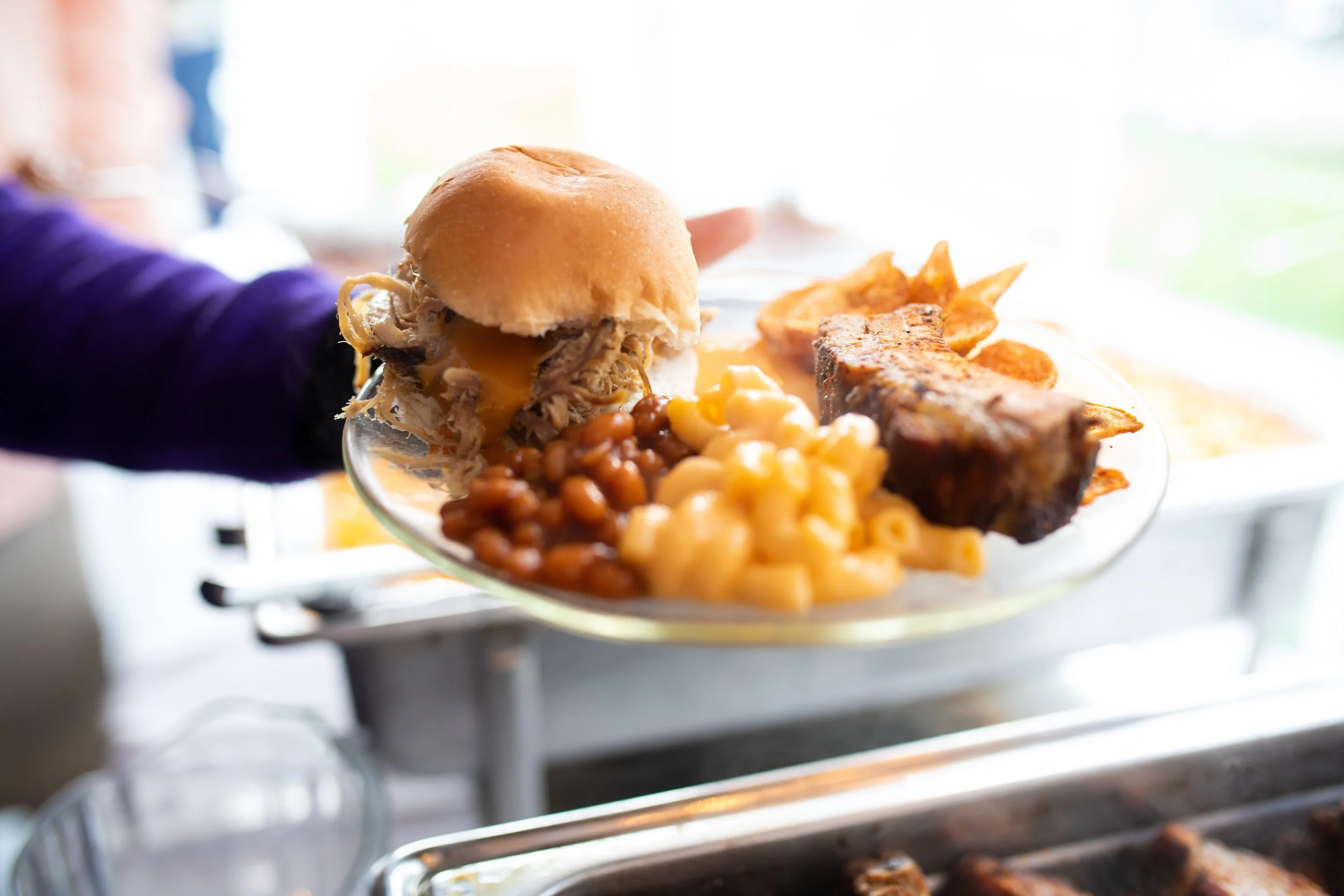 A plate of barbecue food, including pulled pork sandwich, baked beans, macaroni and cheese, ribs, potato chips, and French fries, being handed over.