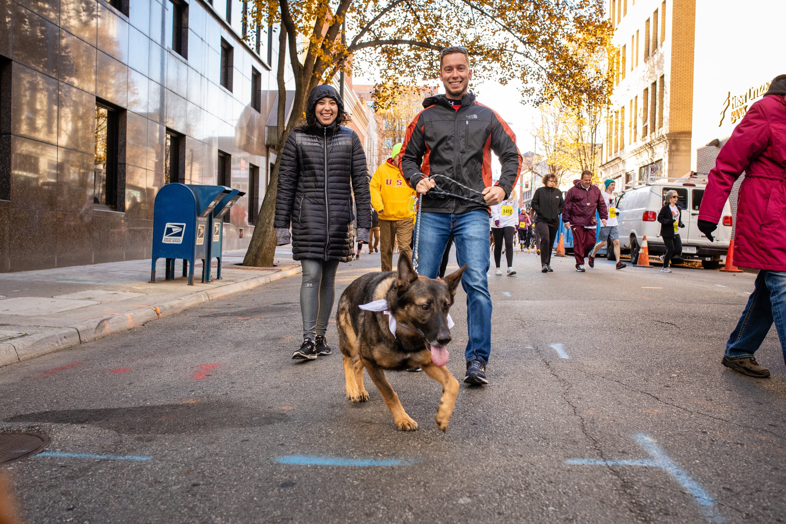 People participating in a street event or race, walking with a dog and smiling, on a city street with buildings, trees, and other participants in the background.