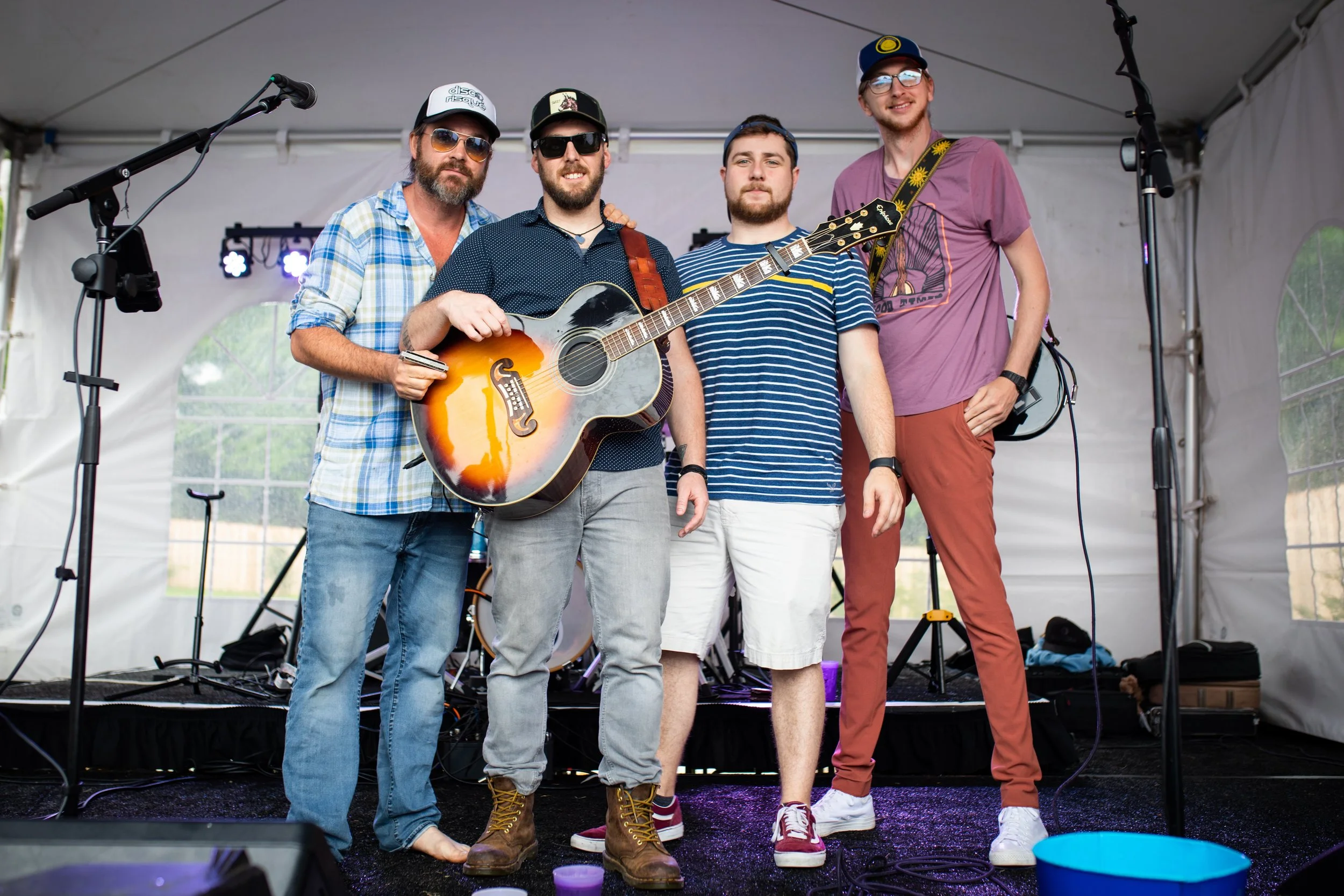 Five men standing on a stage with musical instruments, smiling, in a tent. One holds an acoustic guitar. They are dressed casually with hats and sunglasses.