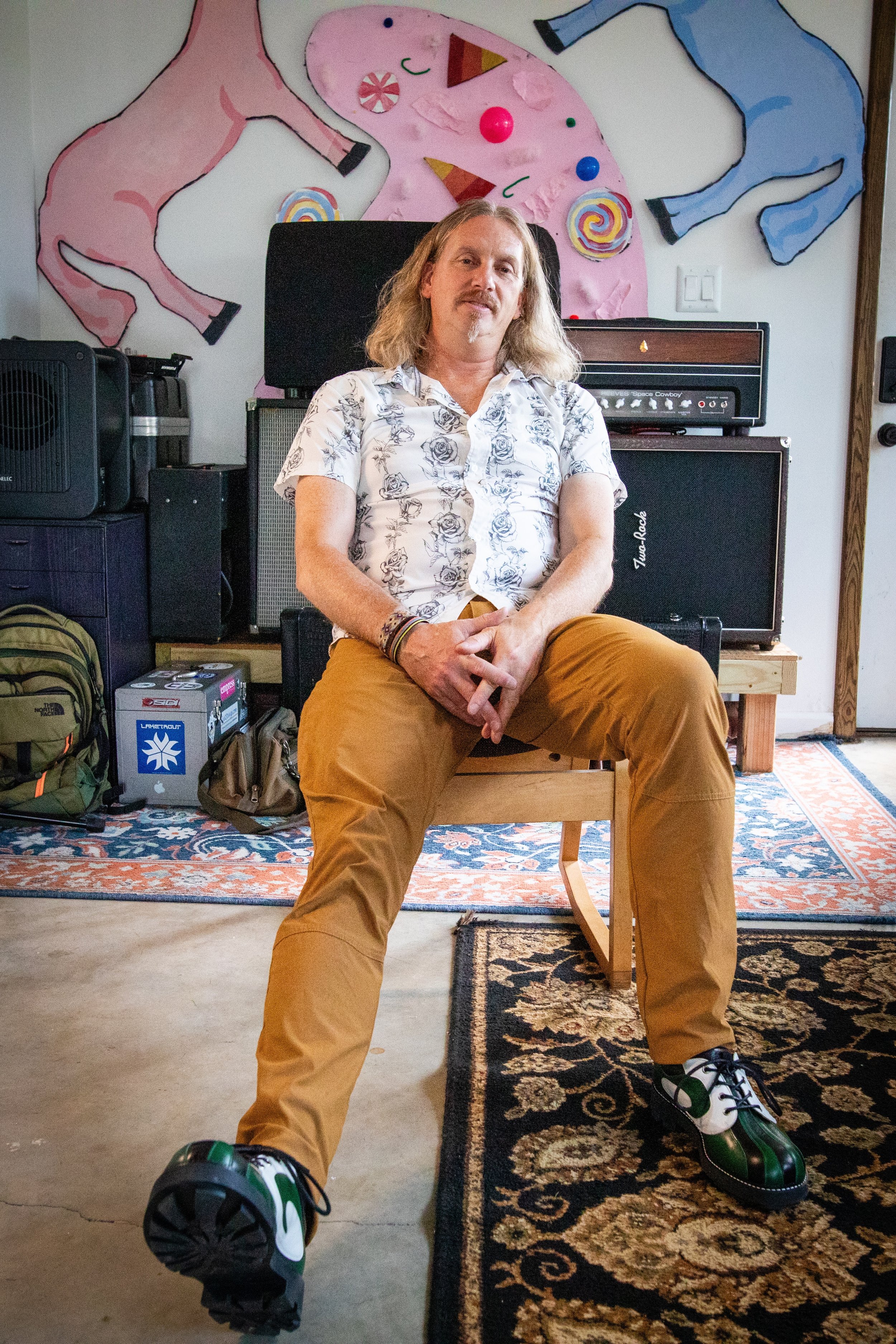 A man with long, wavy hair and a mustache sits on a wooden chair in a room with colorful, abstract animal artwork on the wall behind him. He is wearing a short-sleeved shirt with a floral pattern, tan pants, and green and white shoes. There are music