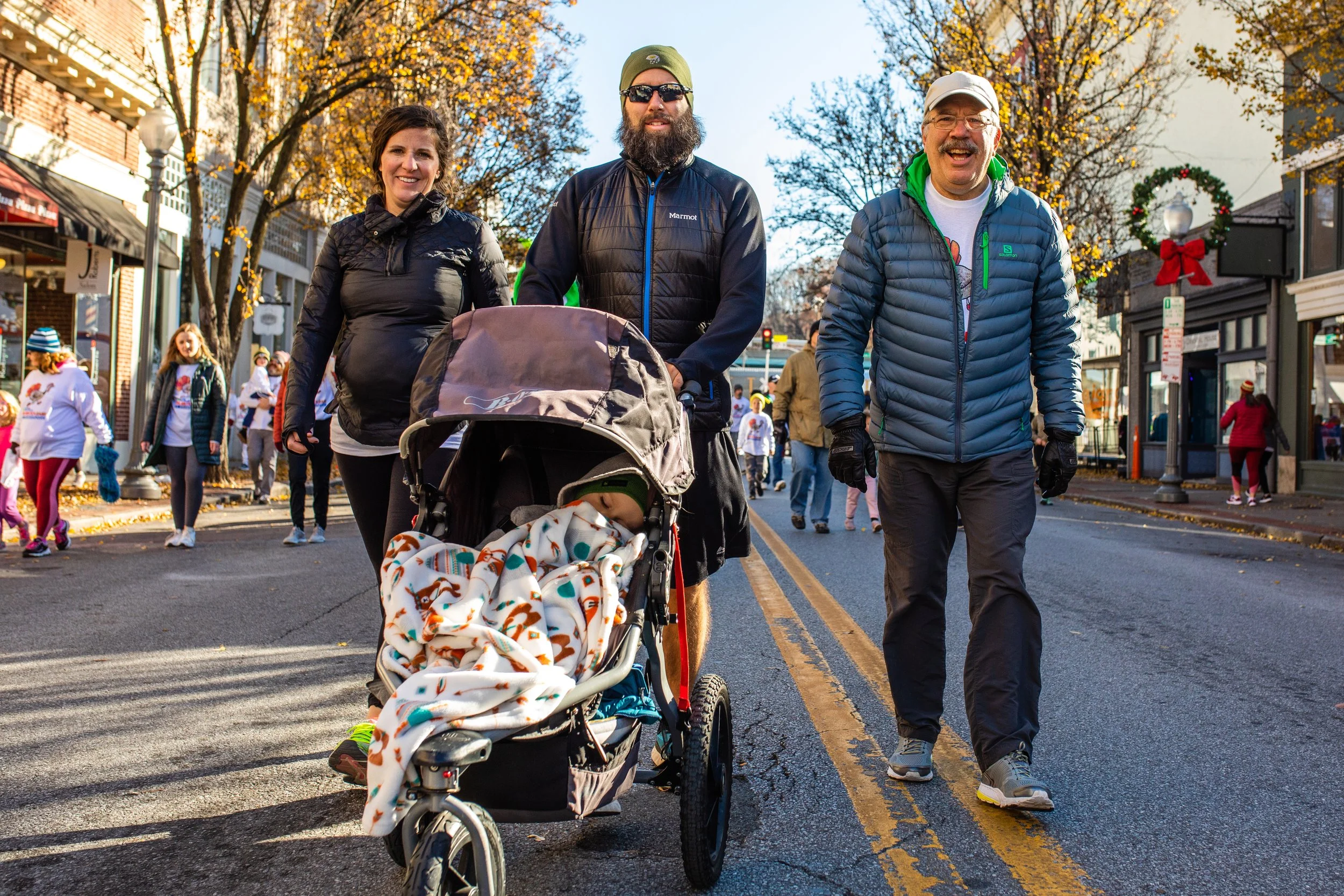 People walking down a city street during a holiday parade or event, with children and spectators in the background, some wearing festive clothing.
