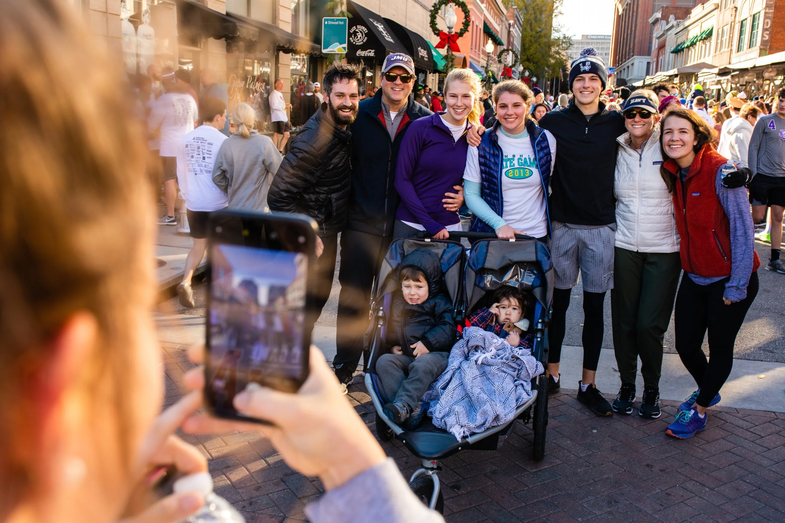 A group of eight people, including two children in a stroller, posing on a busy street with shops and people in the background. One person is taking a photo of the group.
