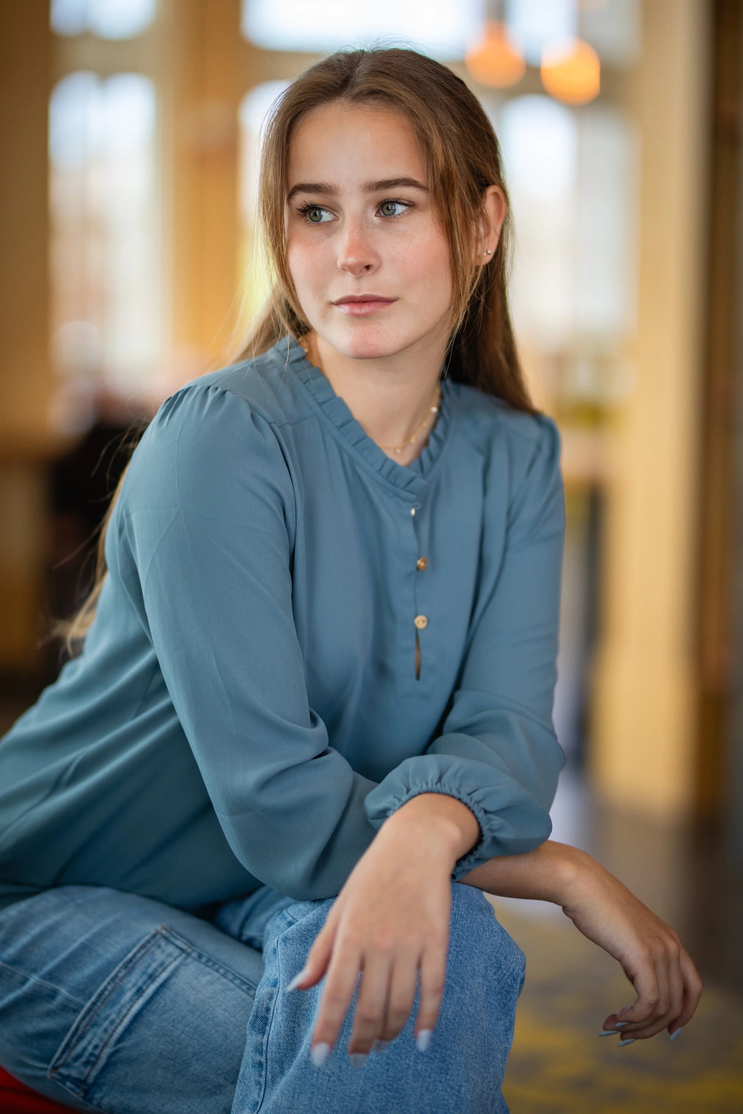 A young woman with long brown hair and blue eyes sitting indoors, wearing a blue blouse and jeans, with a thoughtful expression.