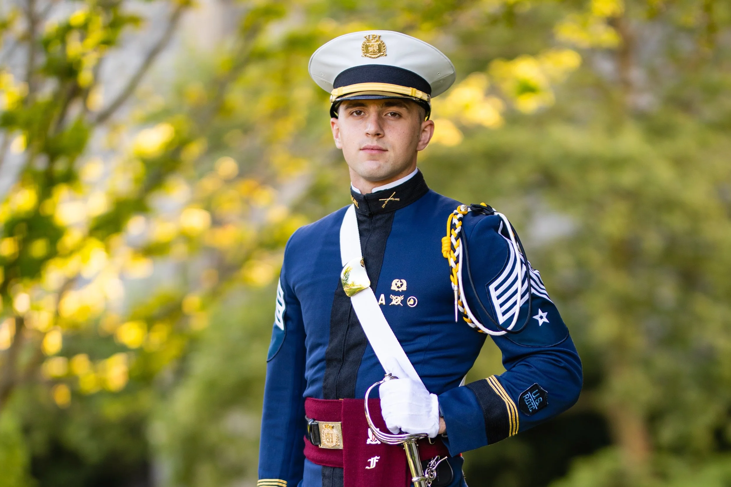 A young man dressed in a military uniform with a white hat, navy blue coat, and white gloves, standing outdoors with green foliage in the background.