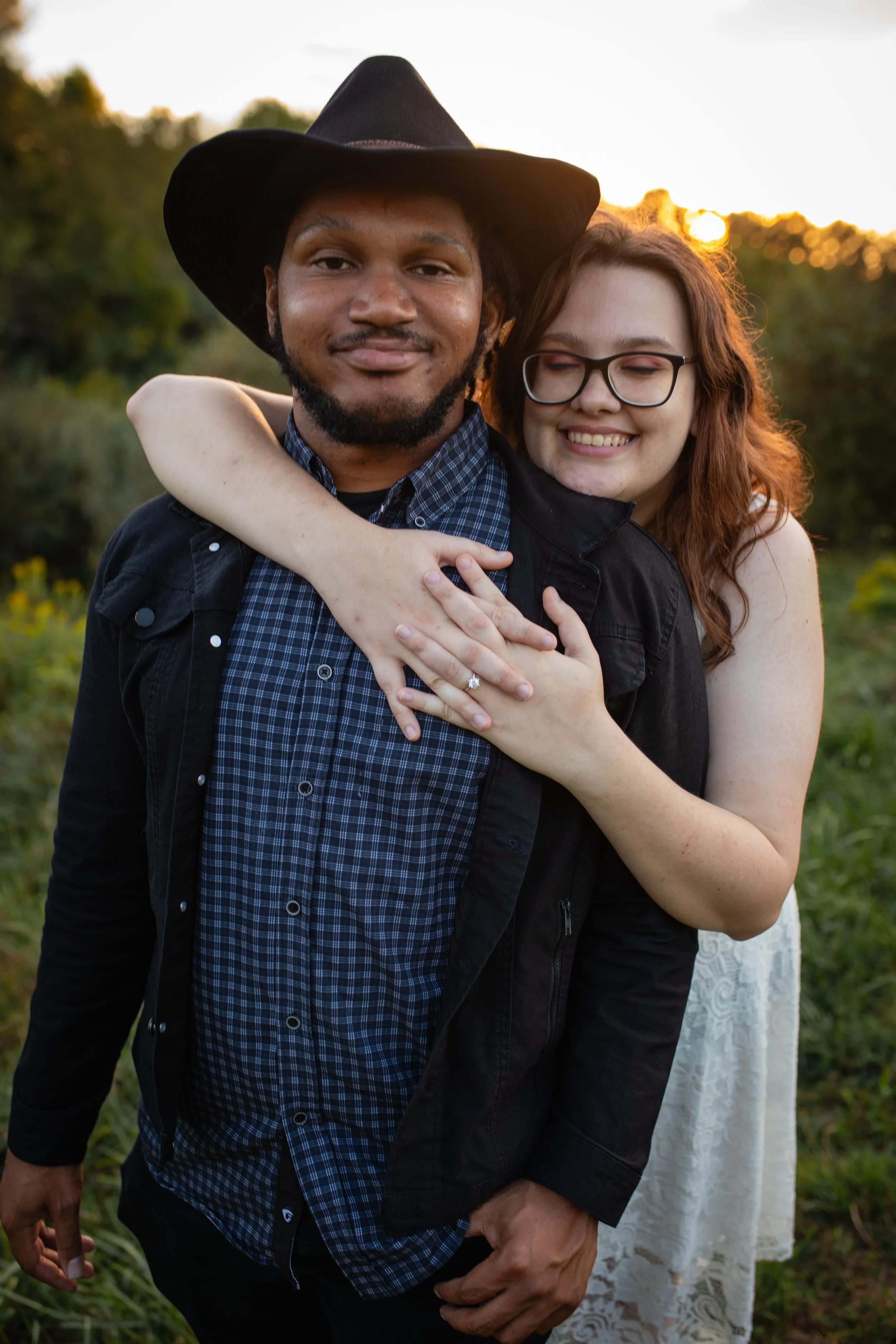 A couple hugging outdoors at sunset, with the woman smiling and the man looking at the camera.