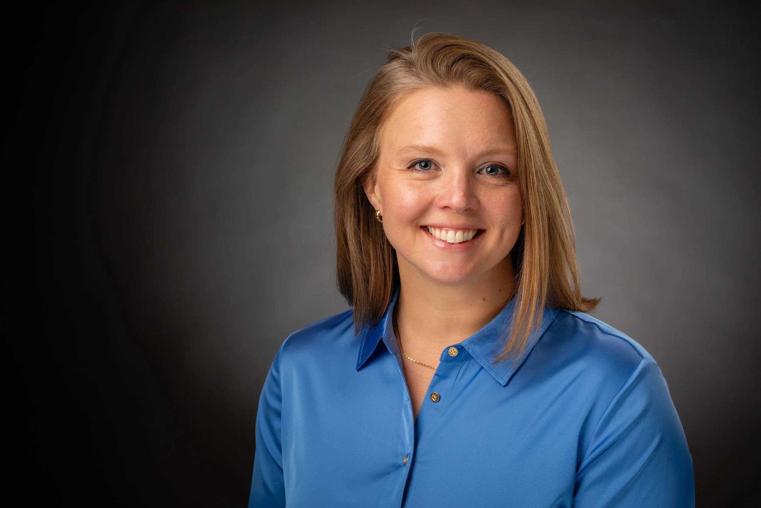 A smiling woman with shoulder-length blonde hair wearing a blue button-up shirt against a dark gray background.