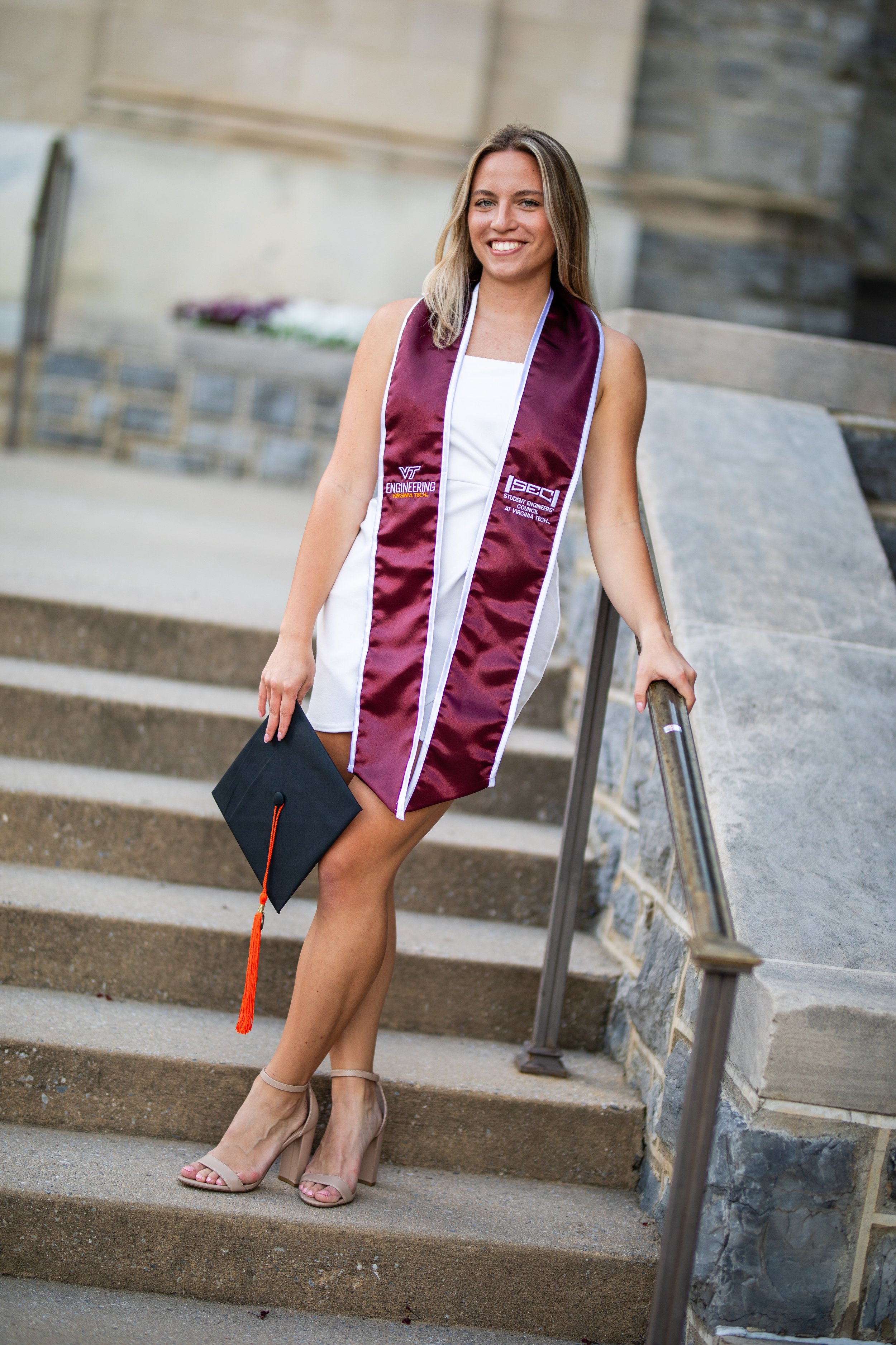 A young woman in a white dress with a Virginia Tech graduation stole, holding a black diploma cover with an orange tassel, standing on outdoor stairs, smiling.