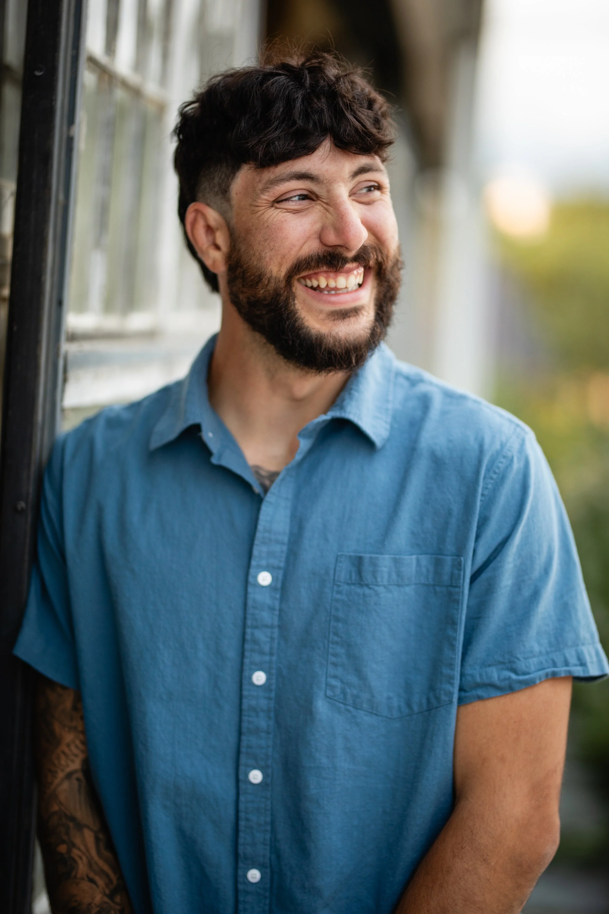 A smiling man with a beard and dark, wavy hair, wearing a blue short-sleeved button-up shirt, standing outdoors near a railing with blurred greenery in the background.