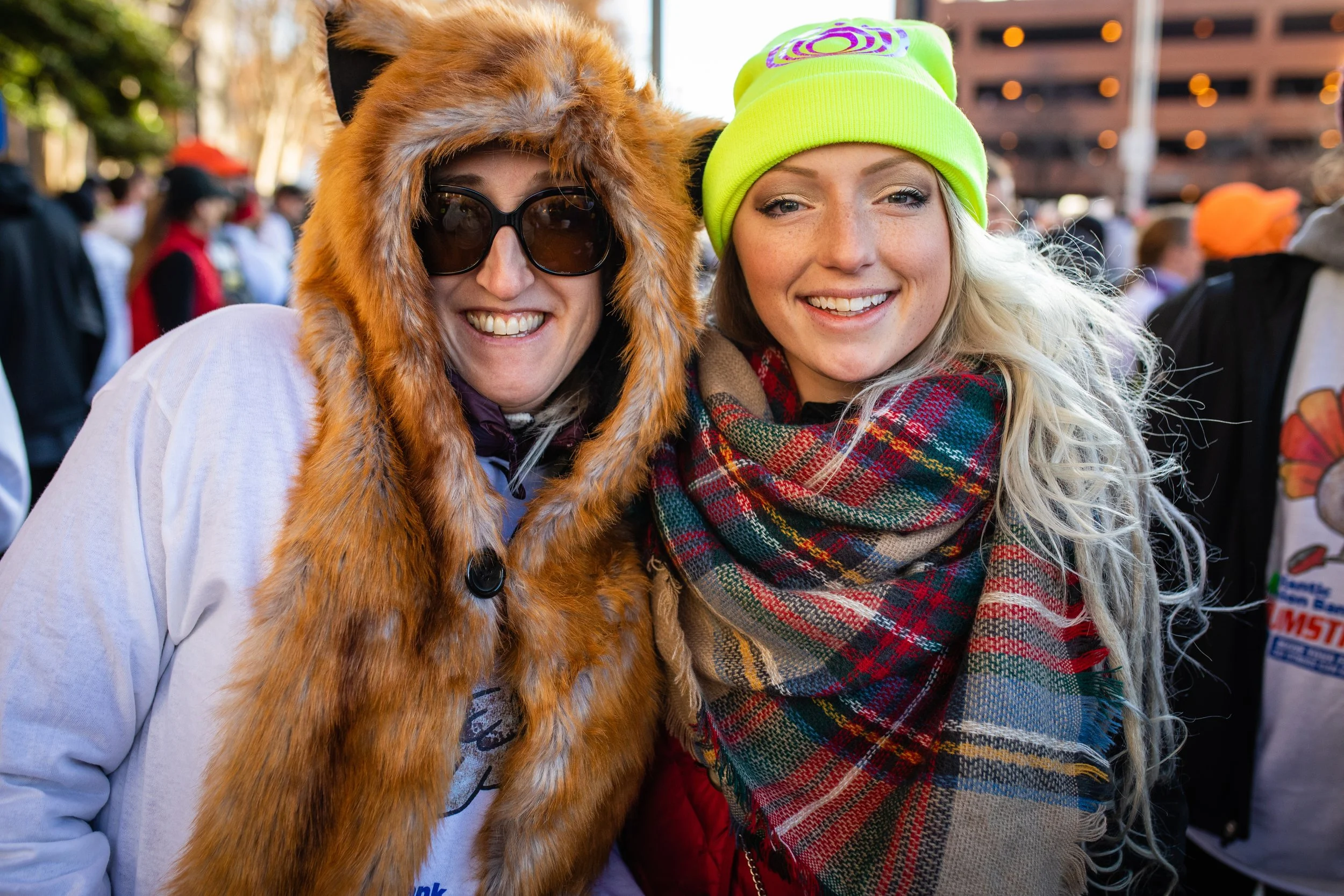 Two women smiling at an outdoor event, one wearing a fox hat and black sunglasses, the other in a neon green beanie and colorful plaid scarf, with a crowd in the background.