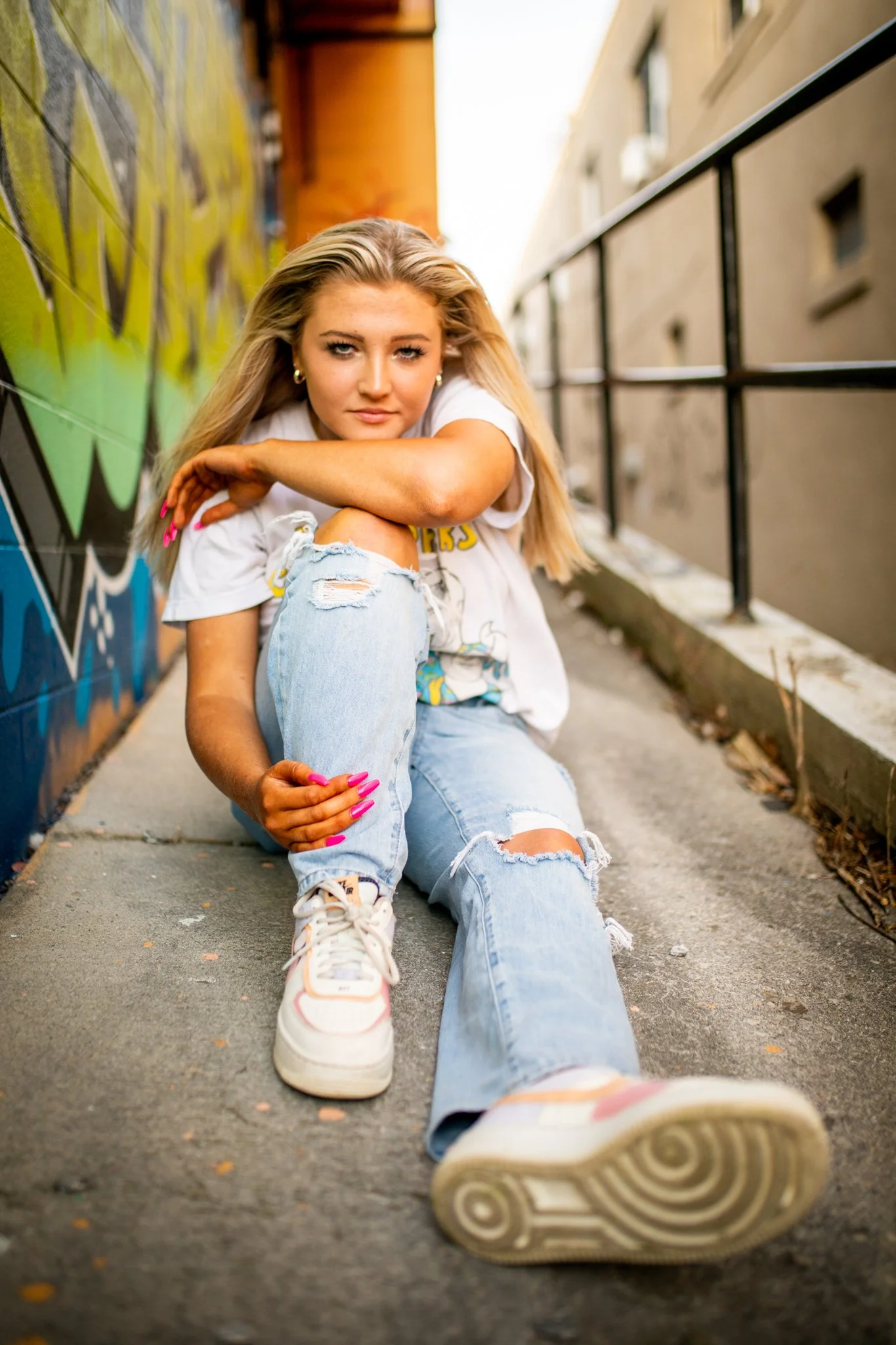 Young woman with long blonde hair sitting on the sidewalk, leaning on her left arm, with graffiti on the wall behind her and buildings on the right.