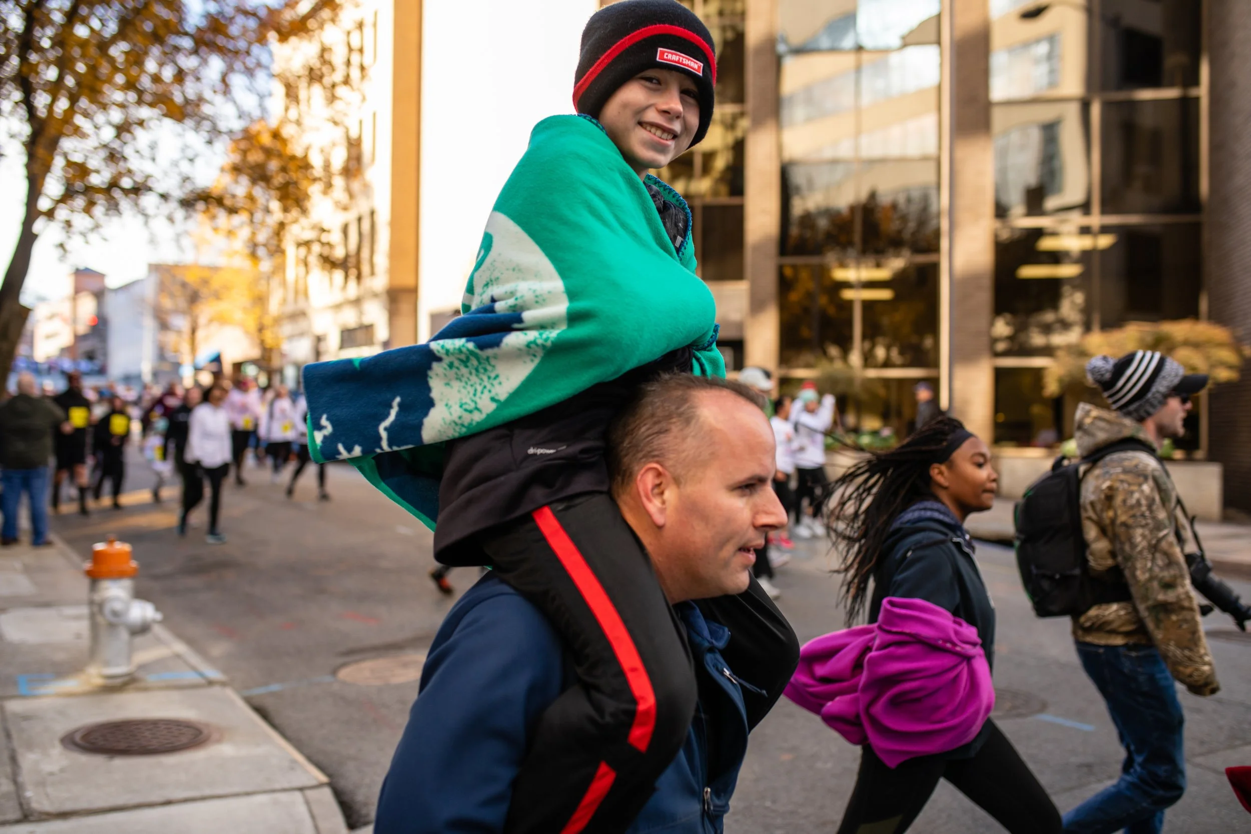 A man carries a boy on his shoulders during a street event or parade, with several people walking and some wearing holiday hats in the background, in an urban setting on a fall day.