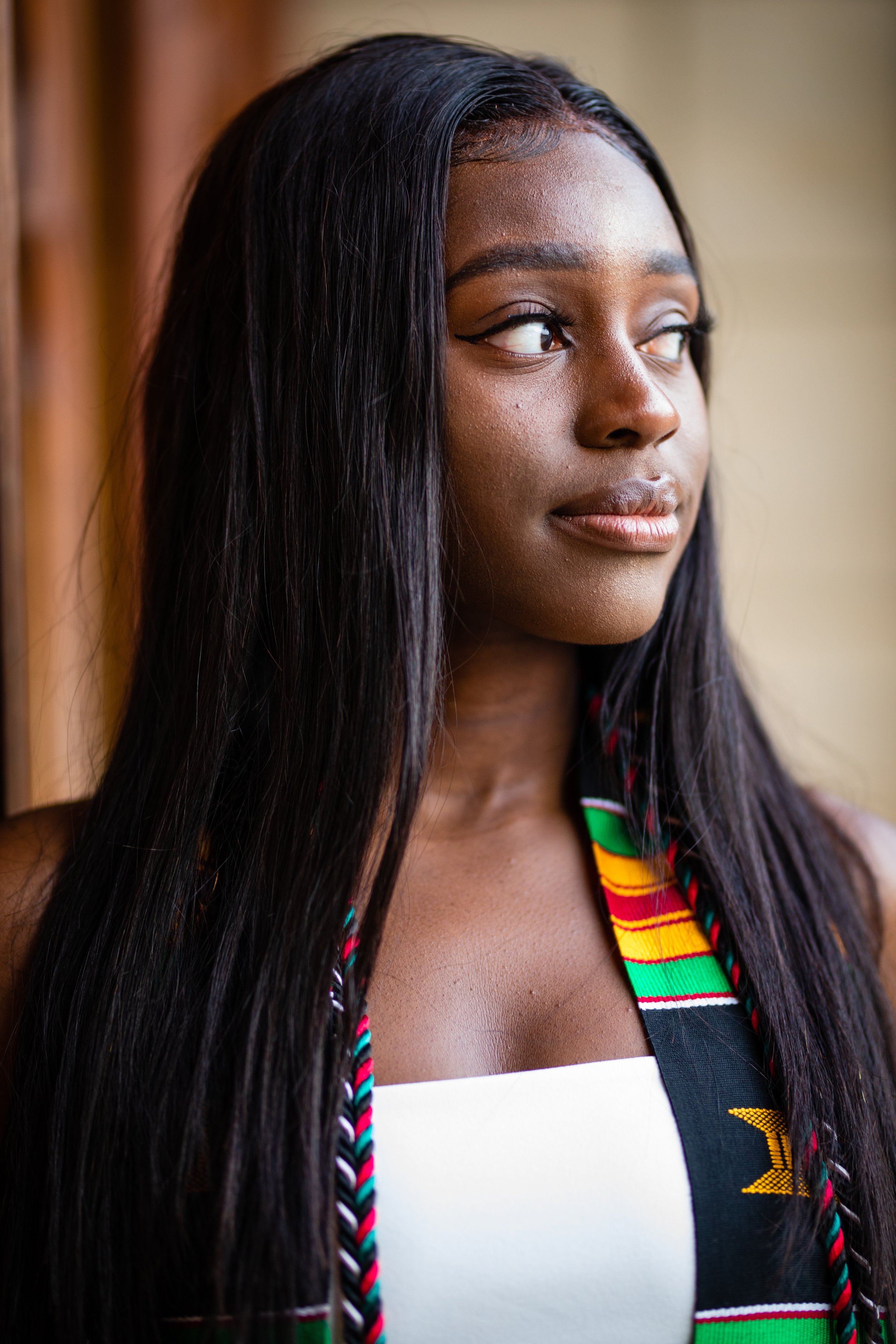 Close-up of a young Black woman with long, straight black hair, wearing traditional African fabric over a white top, looking thoughtfully to the side.
