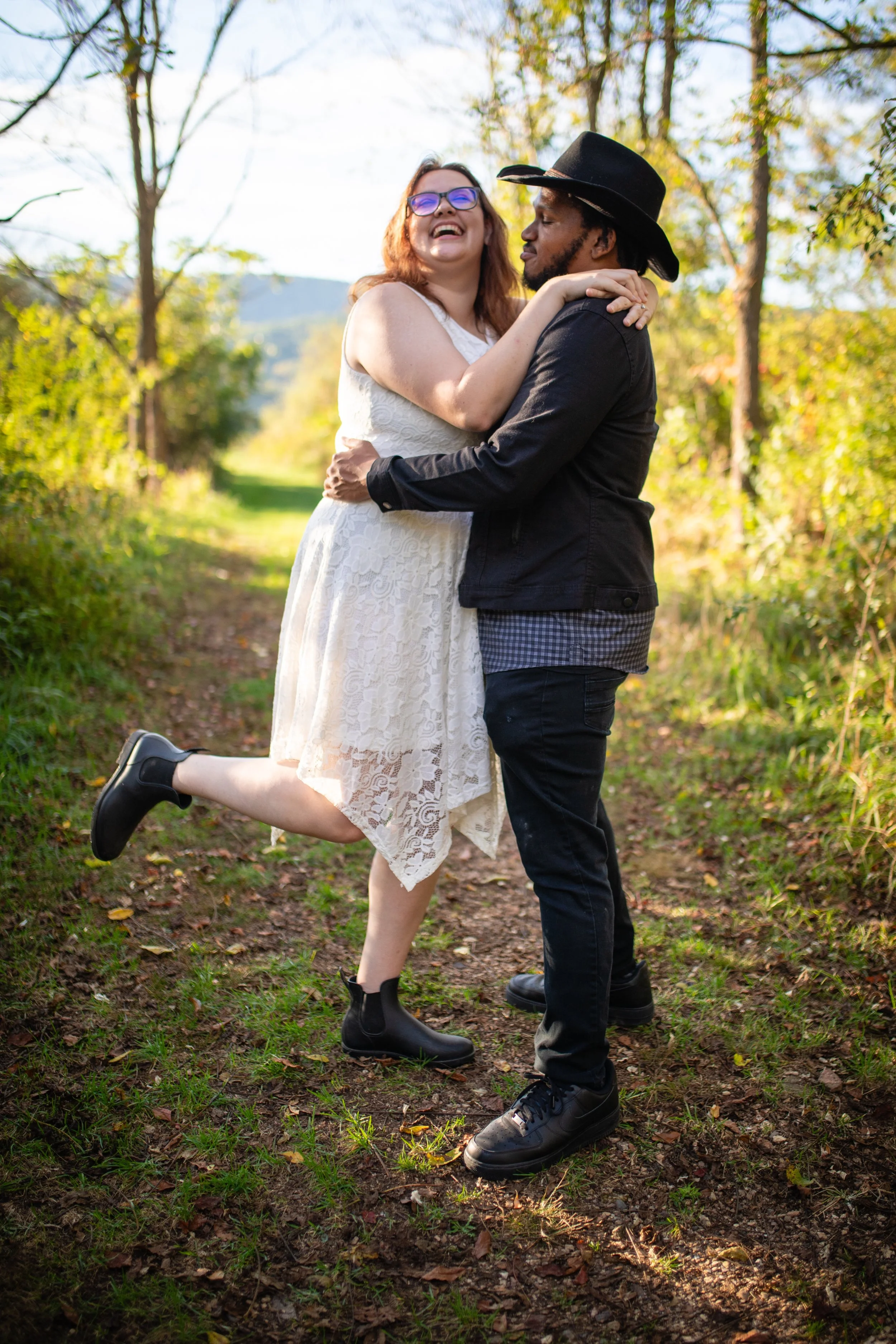 A happy couple embracing outdoors on a dirt trail surrounded by trees and fall foliage. The woman is wearing a white lace dress and black boots, and the man is wearing a black shirt, black pants, and a black hat.