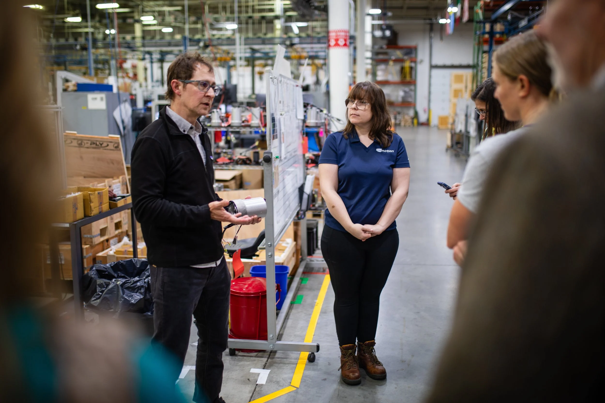A man is holding a cylindrical object and speaking to a group of people in a warehouse or industrial setting, with shelves and equipment in the background.