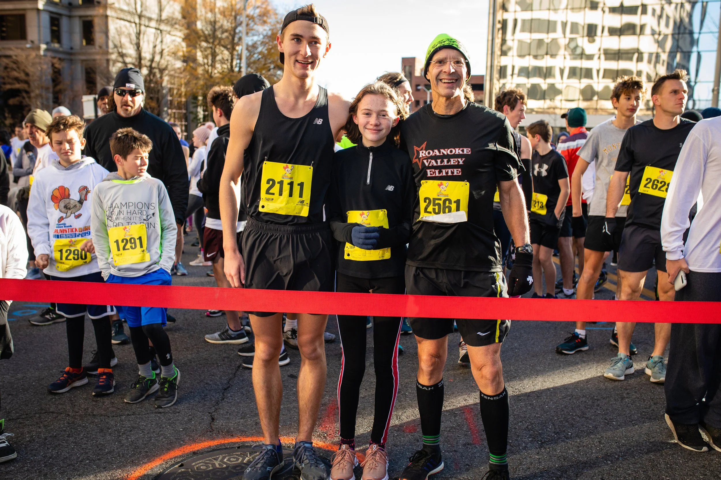 Three runners at the starting line of a marathon race, smiling and posing for a photo, with a crowd of participants behind them.