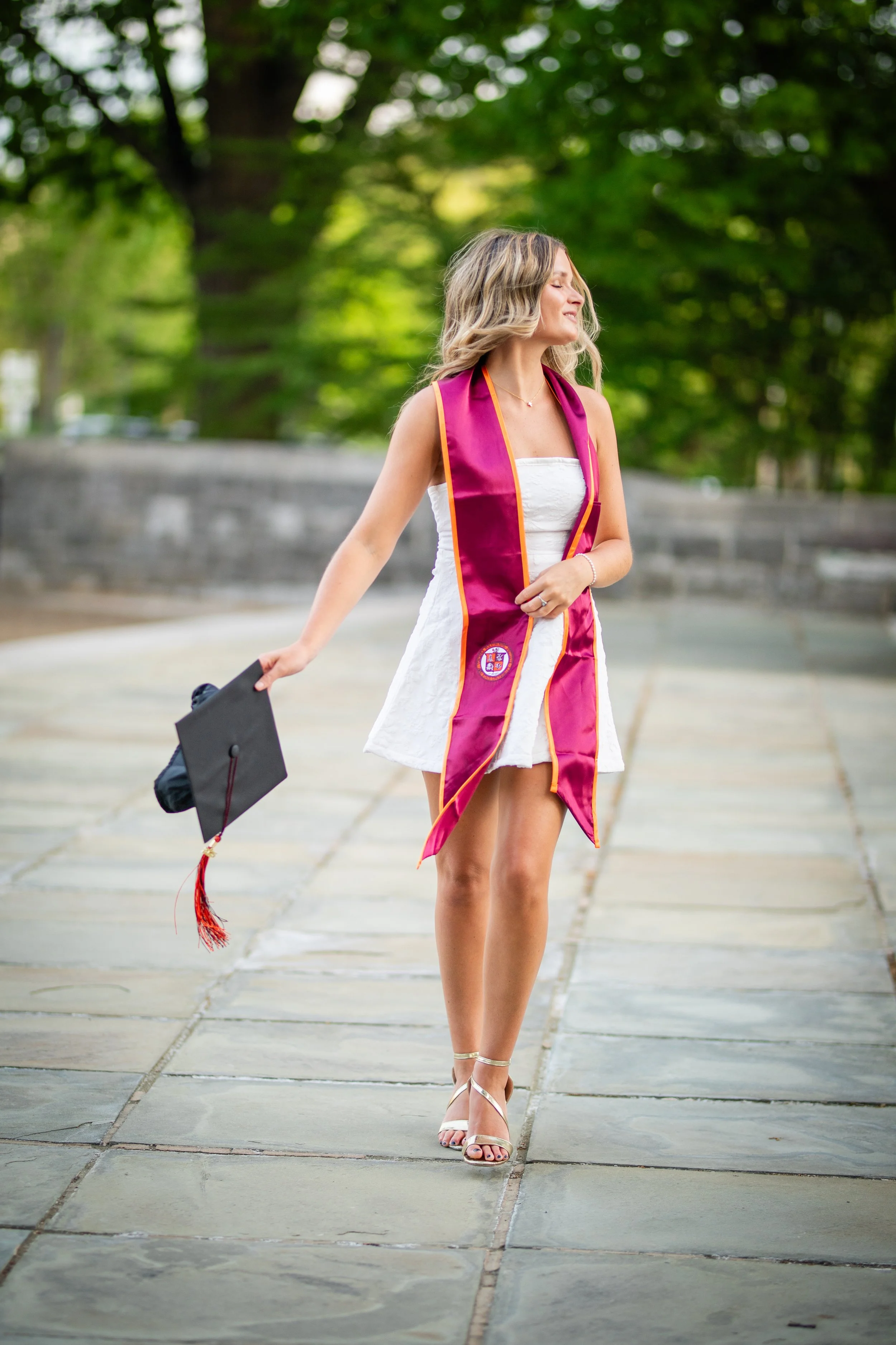 A woman in a white dress and high heels is walking on a sidewalk while wearing a graduation stole. She is holding a graduation cap in her left hand and appears to be celebrating graduation outdoors with green trees in the background.