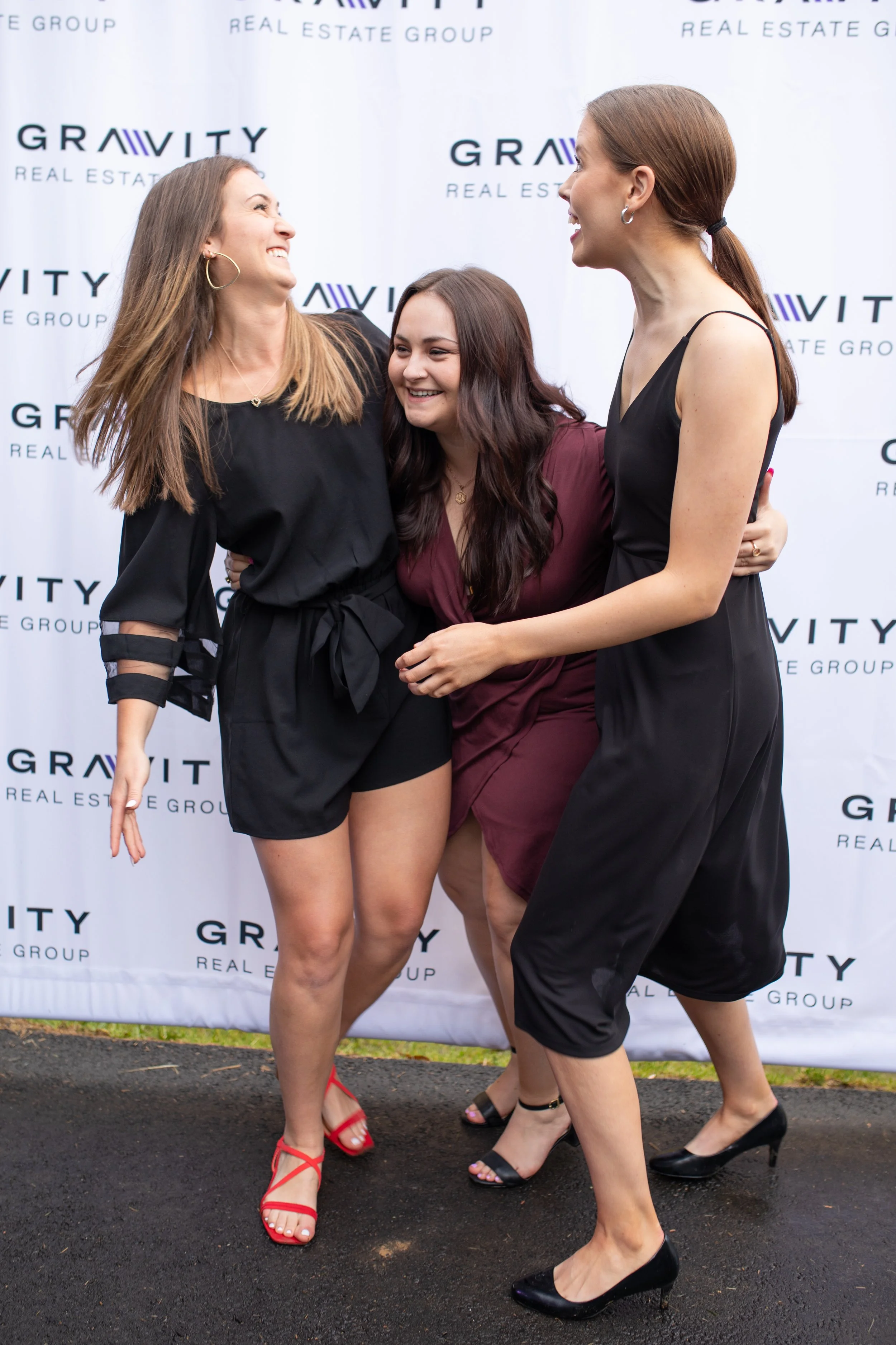 Three women are smiling and hugging each other in front of a backdrop with the logo of Gravity Real Estate Group. They are dressed in black and burgundy dresses and high heels.