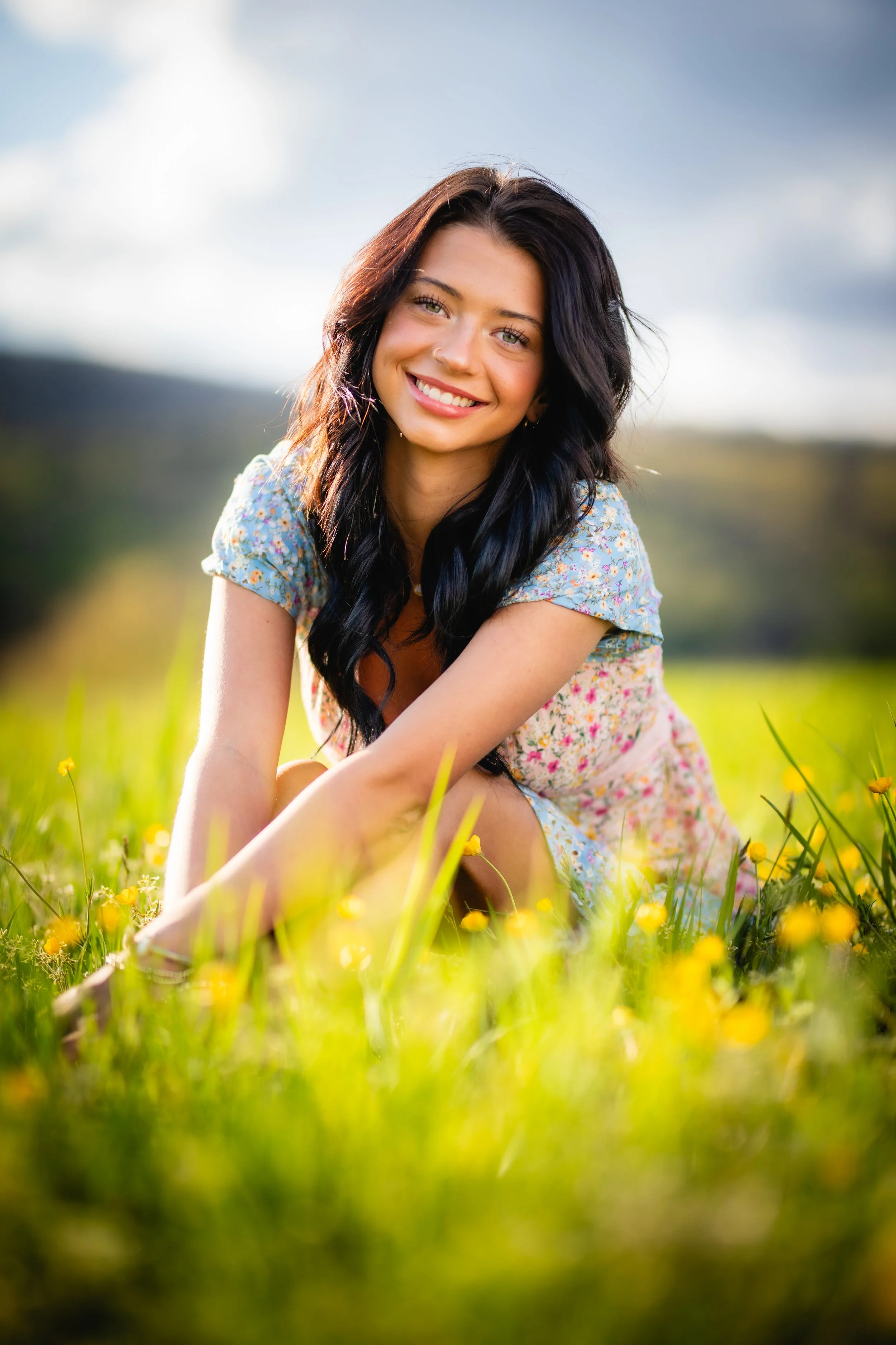 A smiling woman with long black hair wearing a light blue floral dress, sitting in a field of yellow flowers with a blurred green landscape and cloudy sky in the background.
