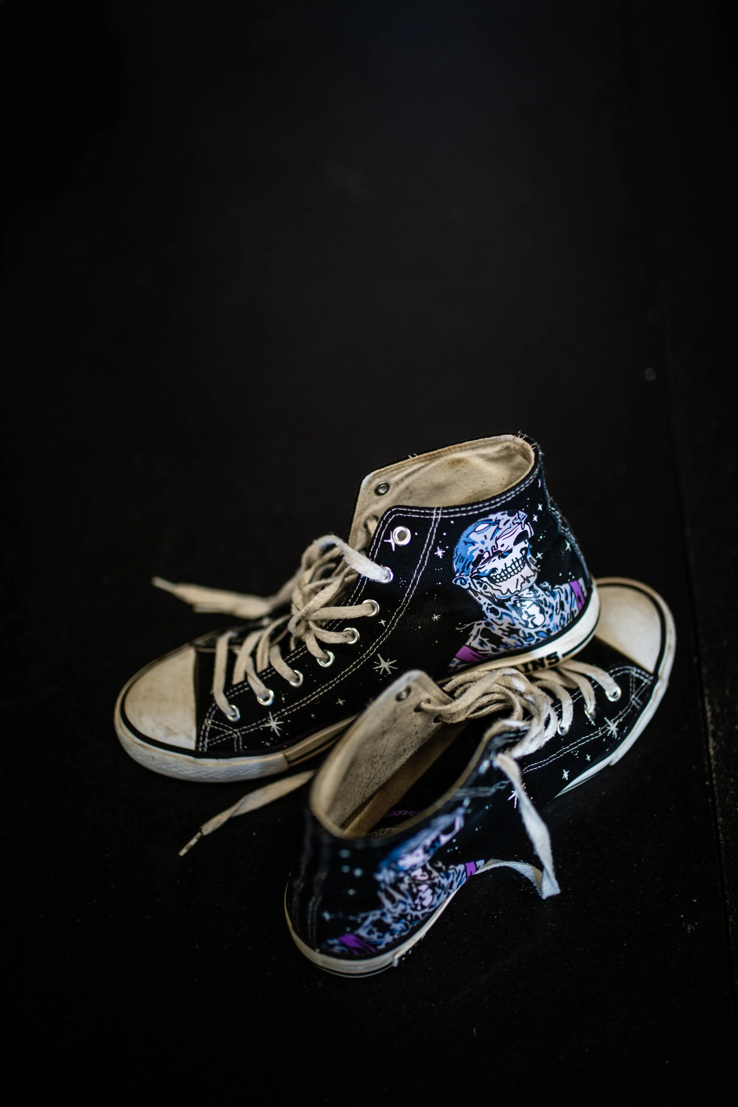 A pair of black high-top sneakers with space-themed design and skeleton graphic, placed on a black surface.