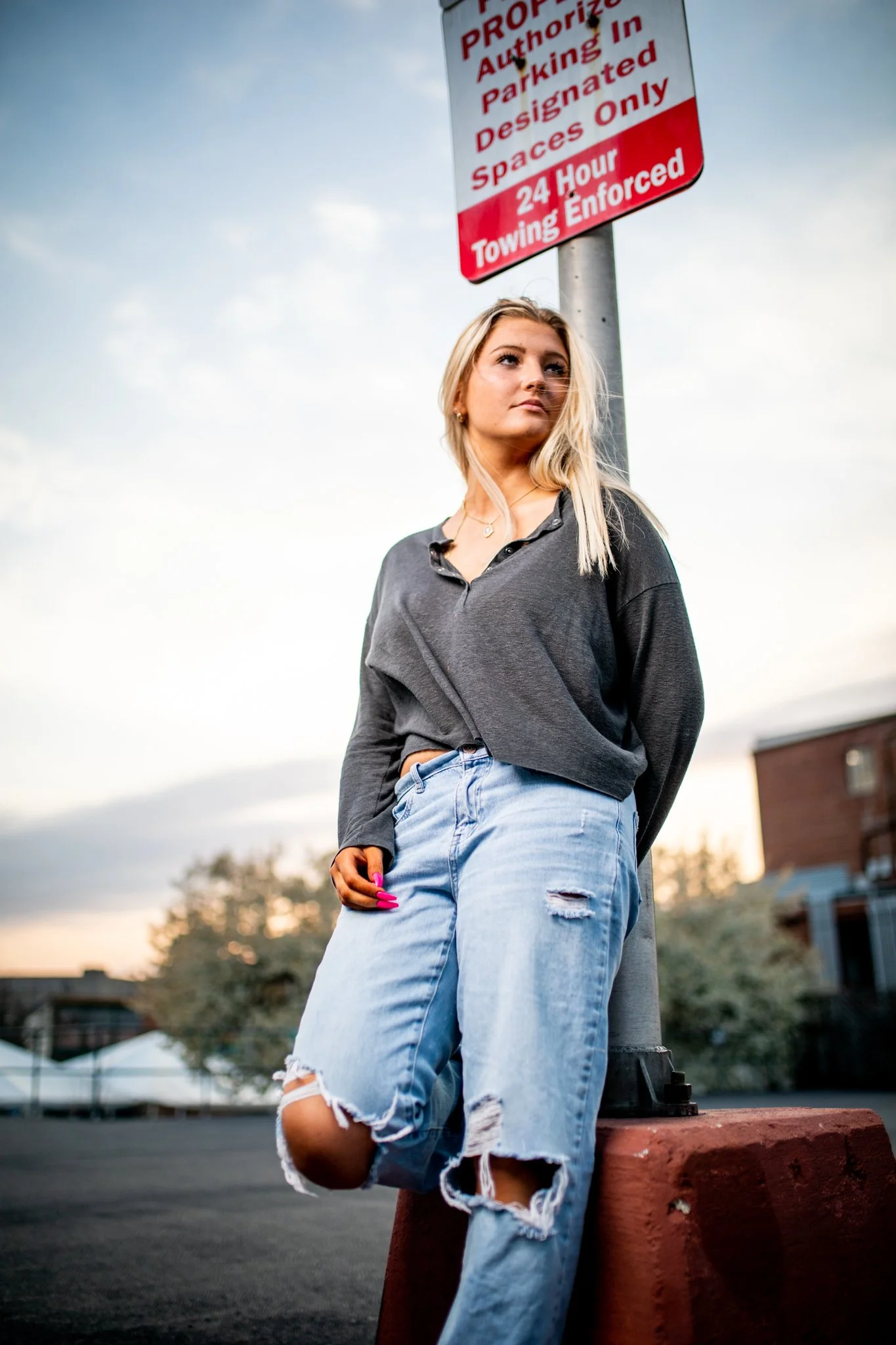 A young woman with long blonde hair wearing a dark gray long-sleeve shirt and ripped jeans standing next to a parking regulation sign in an outdoor parking lot during sunset.